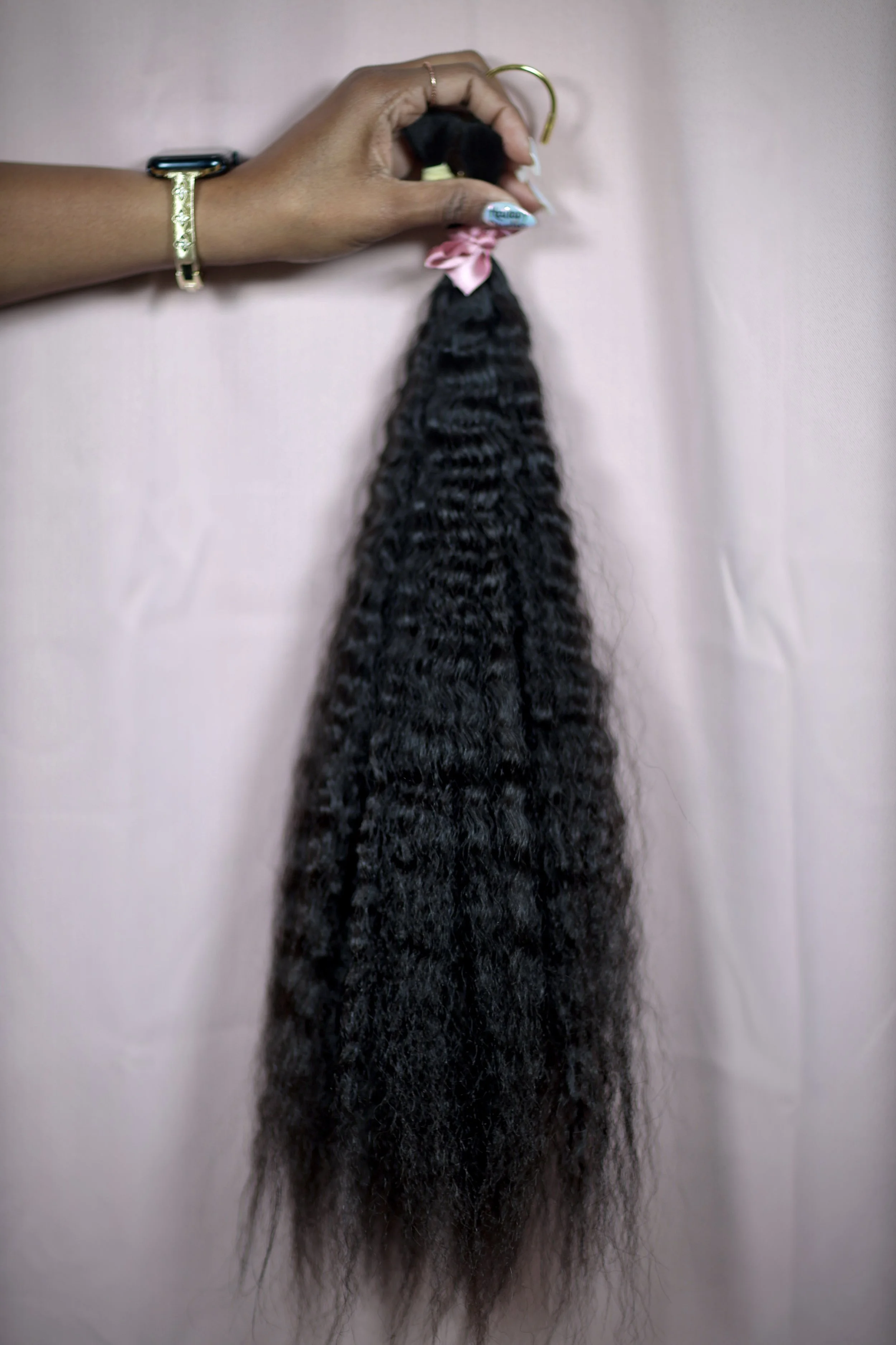 Hand holding a lock of curly black hair tied with a pink ribbon, against a pink background.