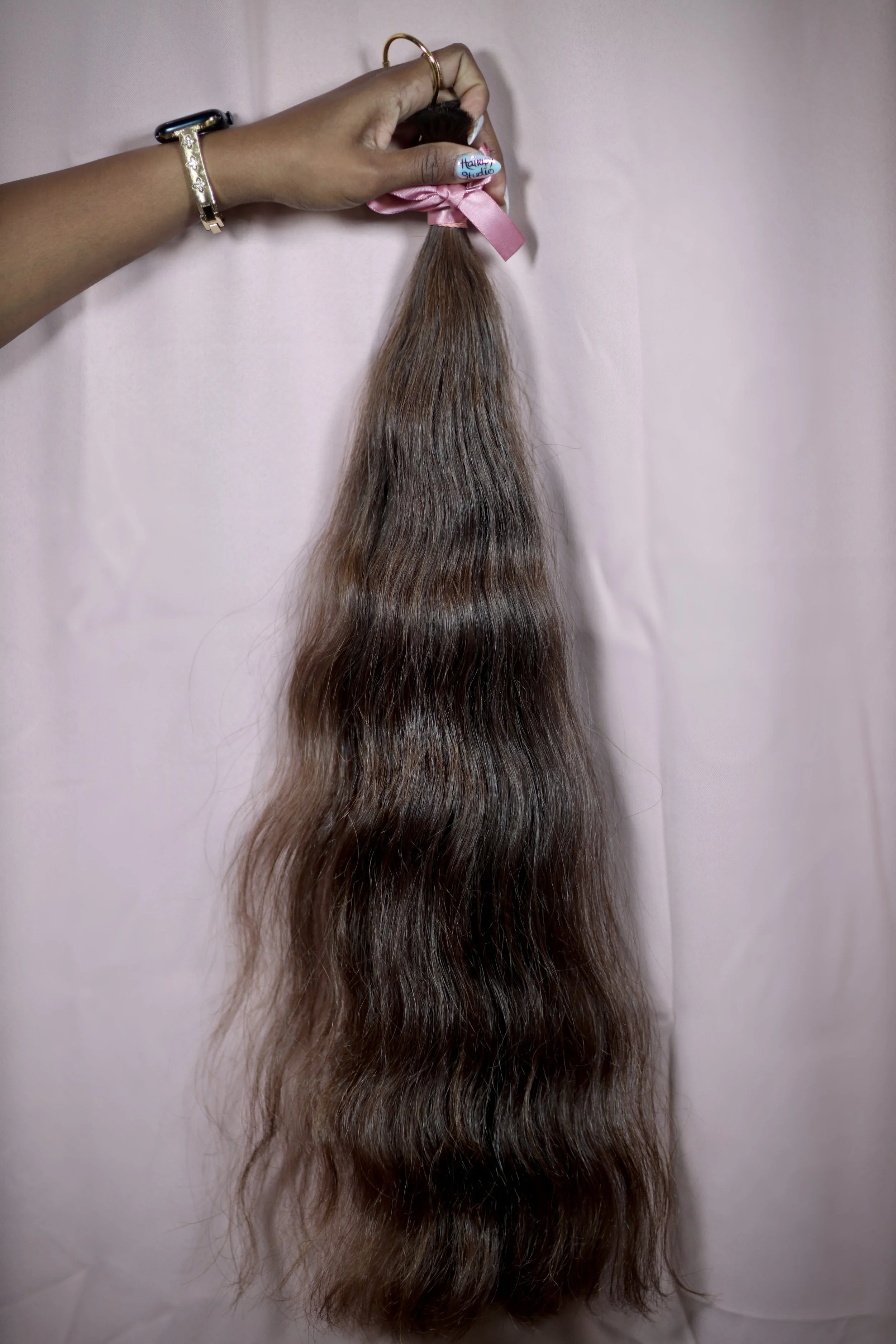 A person's hand holding a bundle of long brown hair tied with a pink ribbon against a pink background.