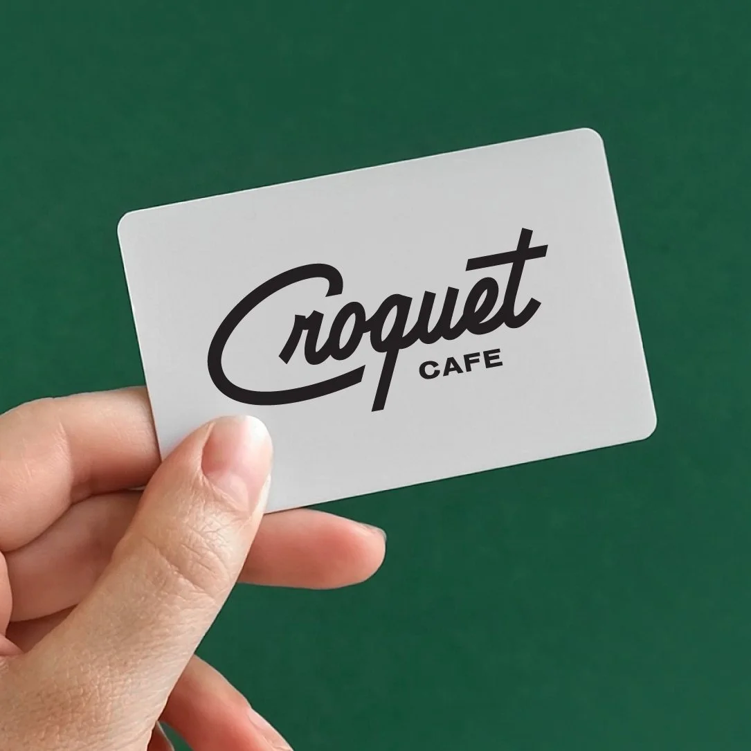 Hand holding a white card with the text 'Croquet Café' against a green background.