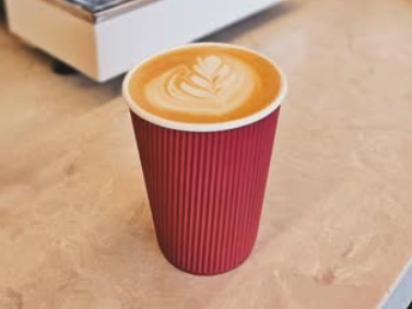A red disposable coffee cup with a latte art heart on top, placed on a beige surface.