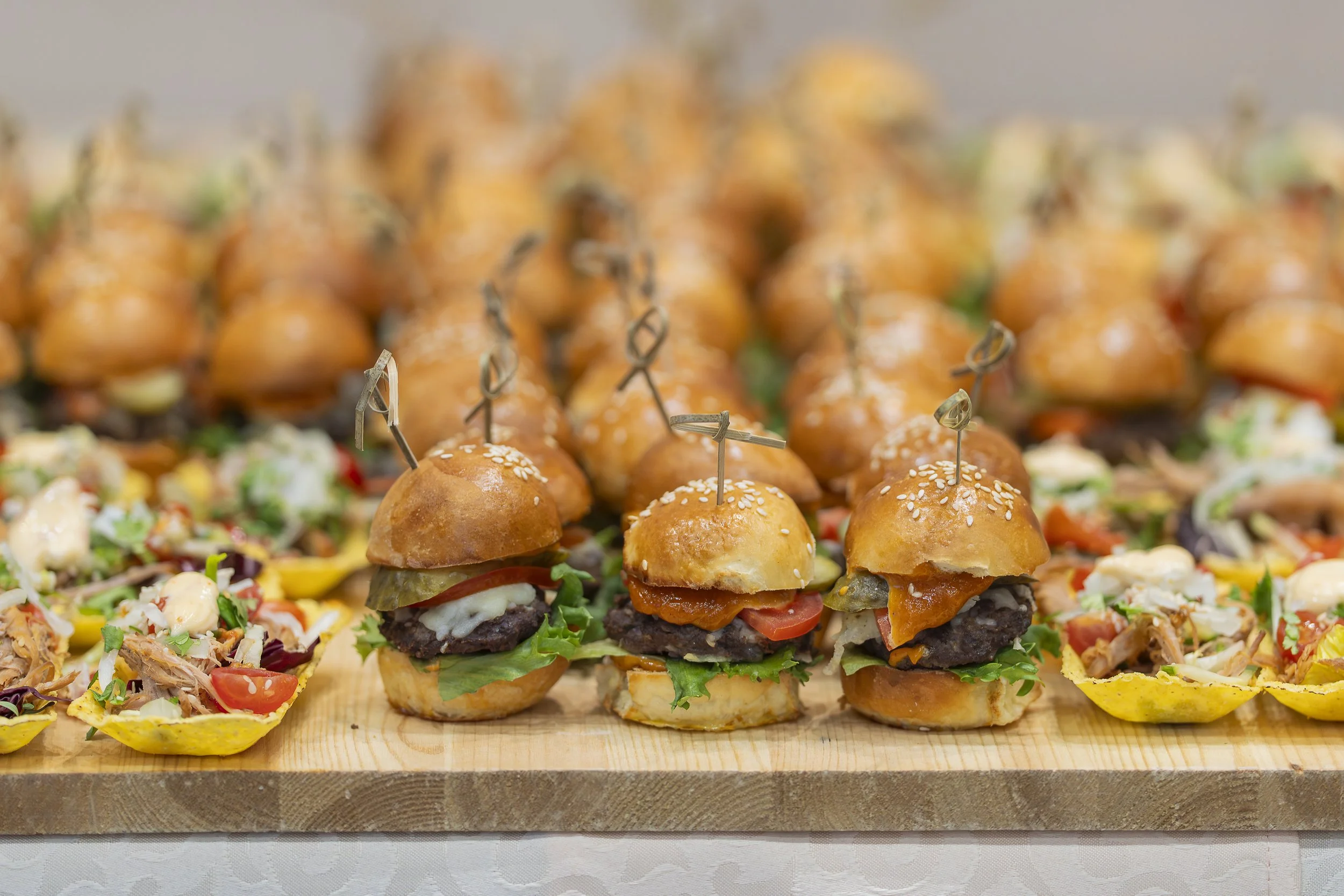 A tray of miniature cheeseburgers on a wooden surface surrounded by small taco shells filled with assorted ingredients like vegetables and sauces.