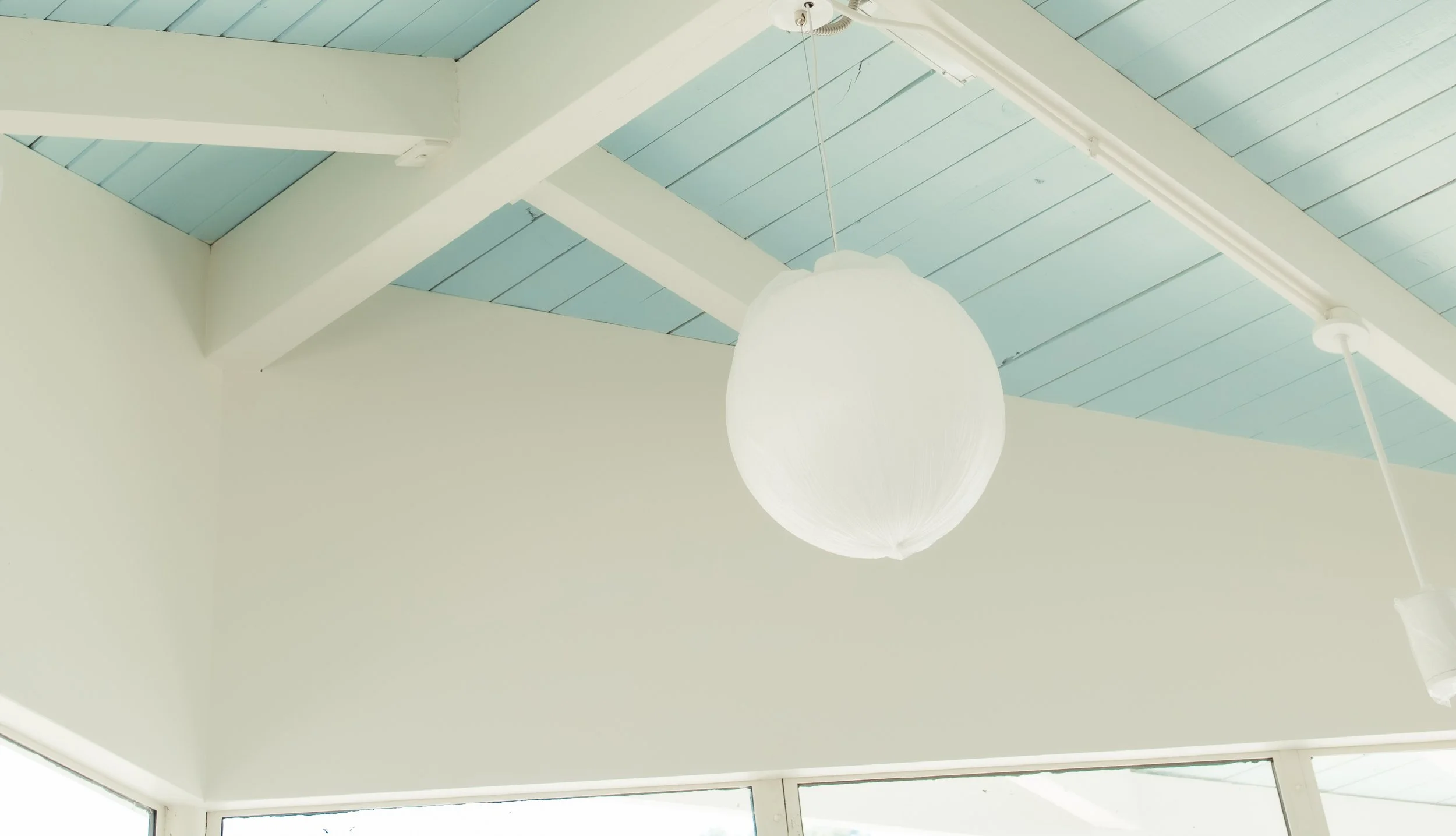 Interior view of a ceiling with white beams and a blue ceiling panel, featuring a large round white paper lantern light fixture hanging from a cord.