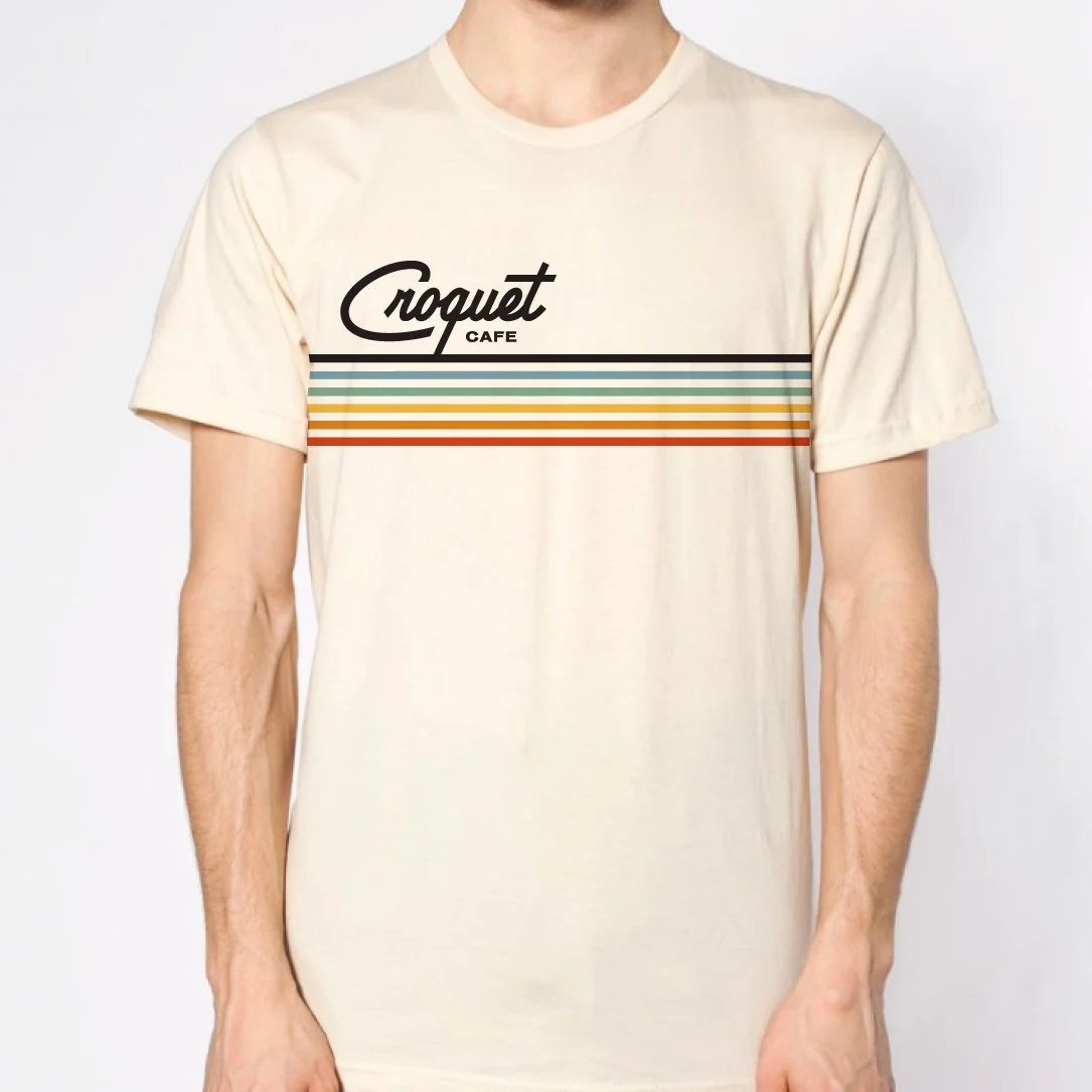 Cream-colored T-shirt with 'Croquet Cafe' logo and multicolored horizontal stripes.