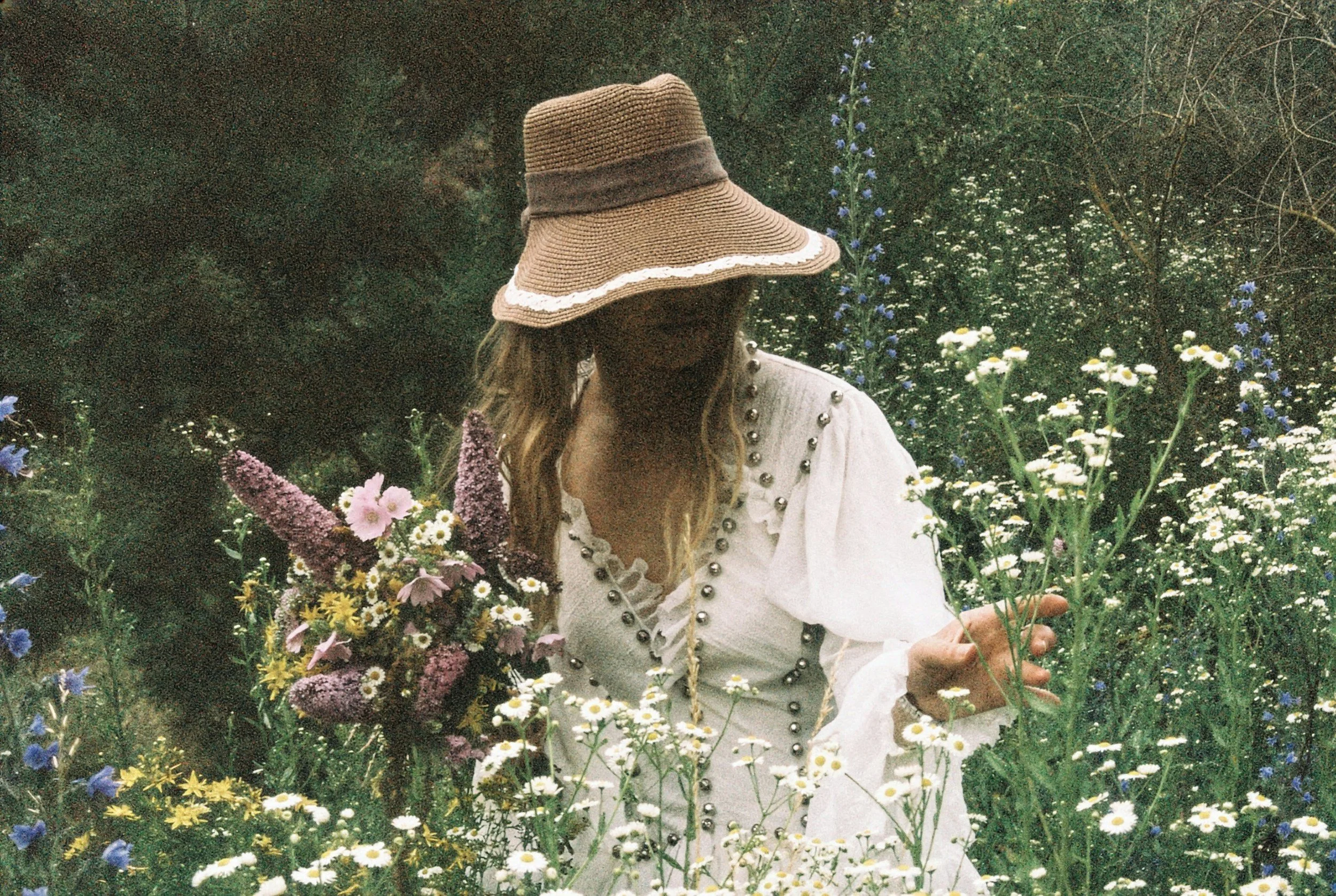Person wearing a wide-brimmed straw hat surrounded by various wildflowers in a garden.