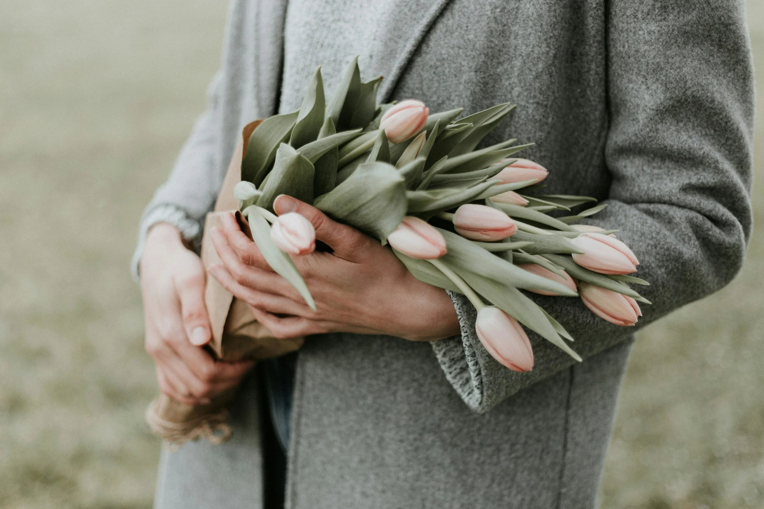 Person holding a bouquet of pink tulips with green leaves, wearing a gray coat.