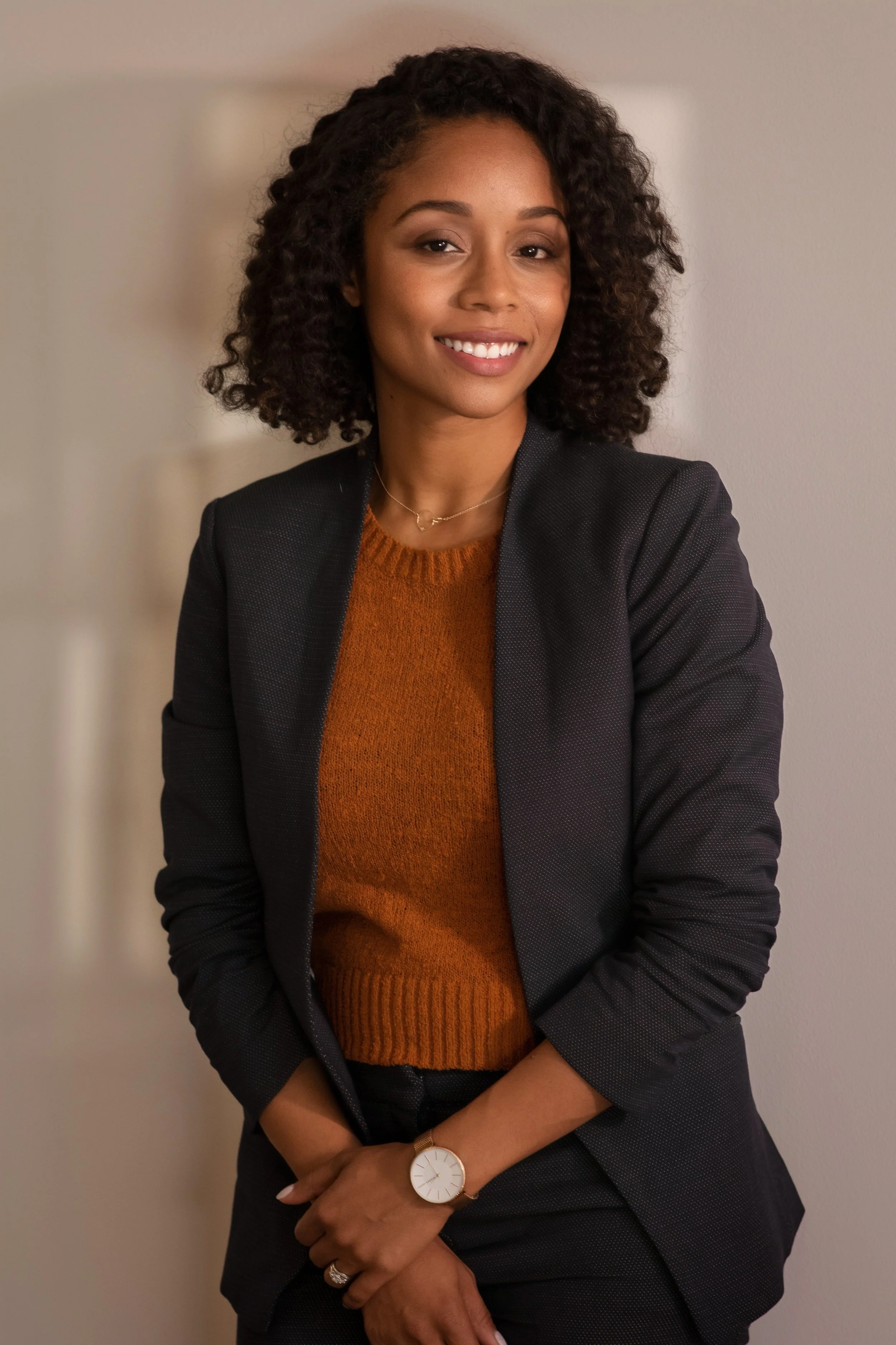 A woman with curly black hair smiling, wearing a black blazer over a rust-colored sweater, a watch, and a ring, standing against a plain wall.