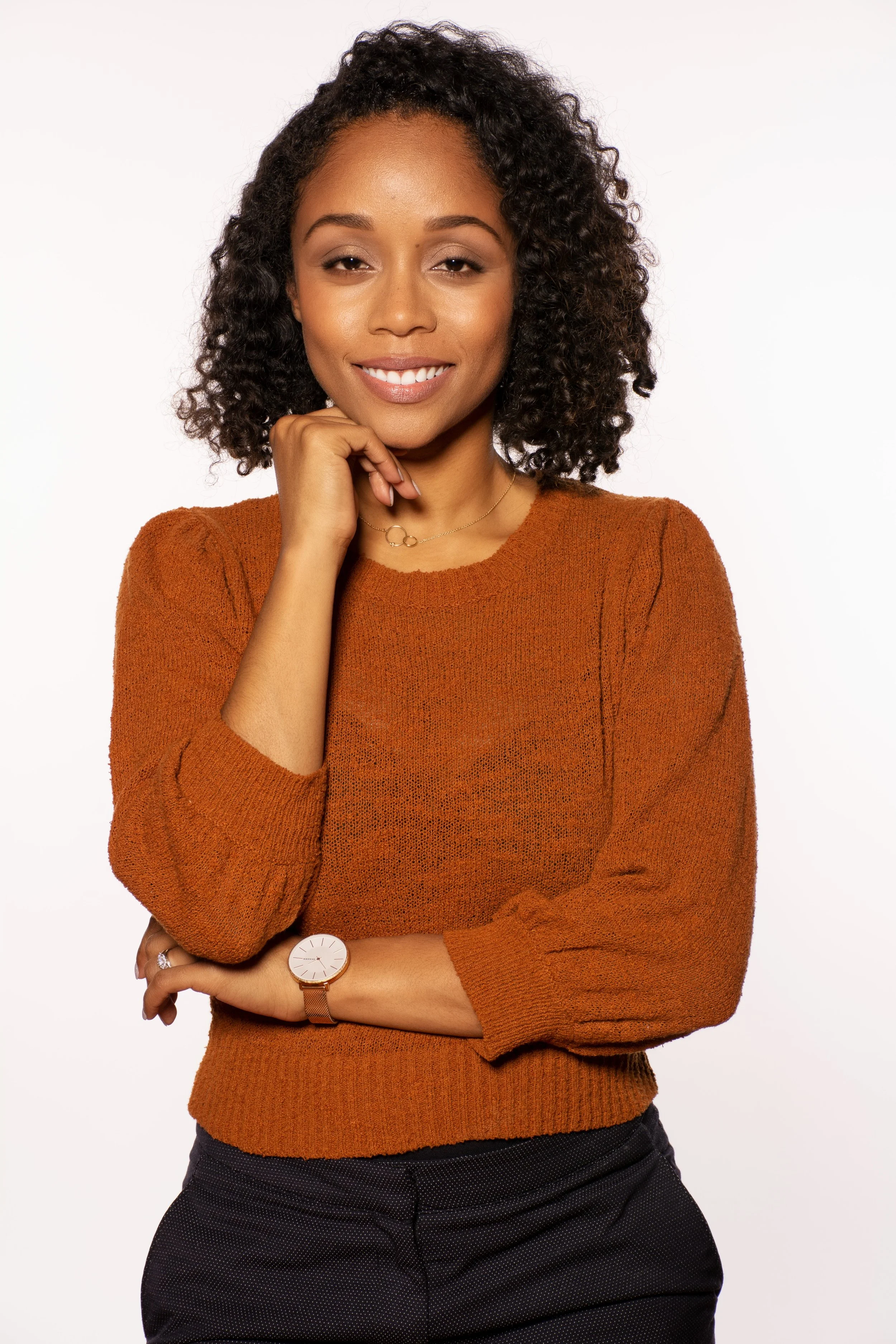 A woman with medium skin tone, curly black hair, wearing a rust-colored sweater, a gold necklace, a watch, and a ring, standing with her hand under her chin and smiling at the camera against a plain white background.