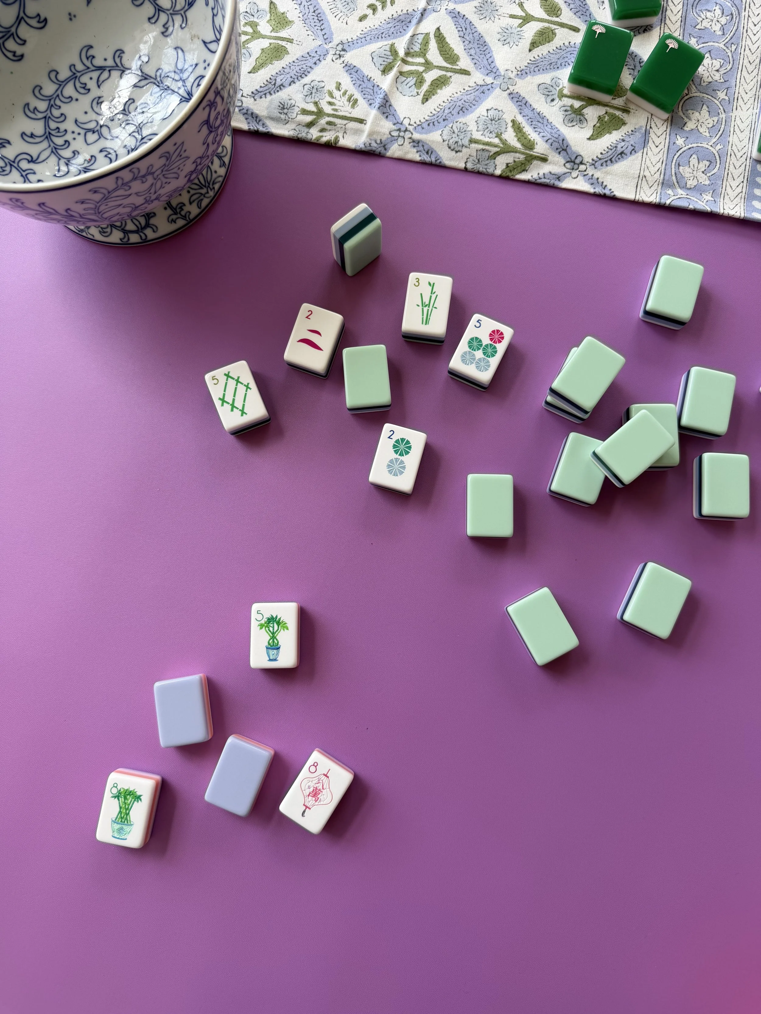 Colorful Mahjong tiles scattered on a pink surface, with some stacked and others lying flat. A decorative bowl and a patterned cloth are partially visible in the background.