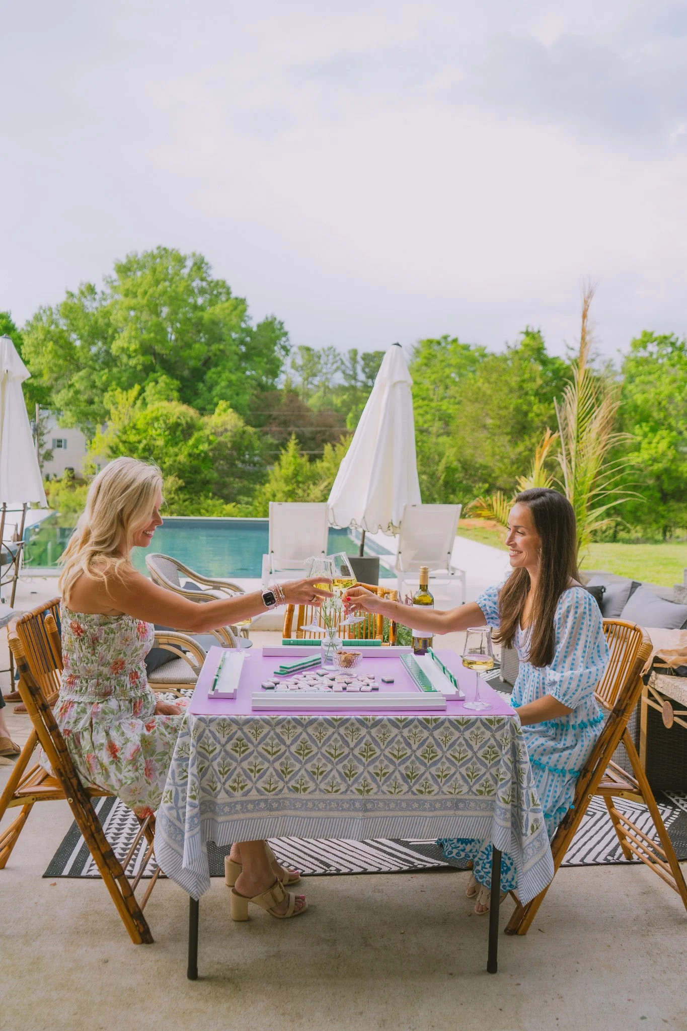 Two women sitting at a table outdoors, playing a board game, and clinking glasses. The scene includes a pool, white umbrellas, and lush green trees in the background.