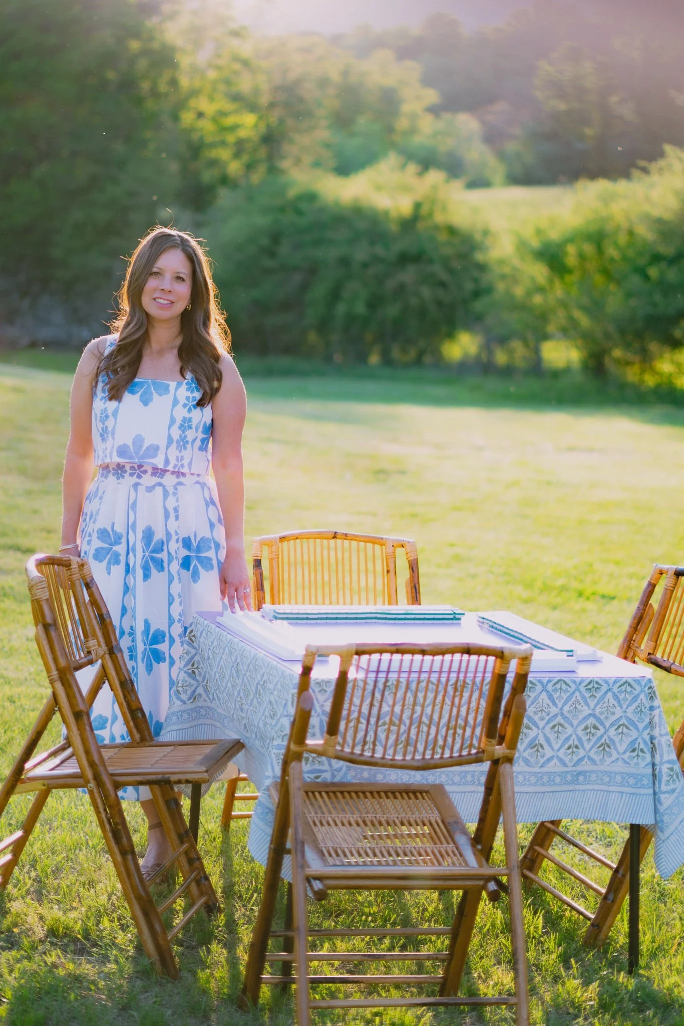 Andrea, the founder of Hill City Mahj, in a blue and white floral dress, stands near a table with stacked notebooks and surrounded by wooden chairs, with lush green trees and a grassy area in the background.