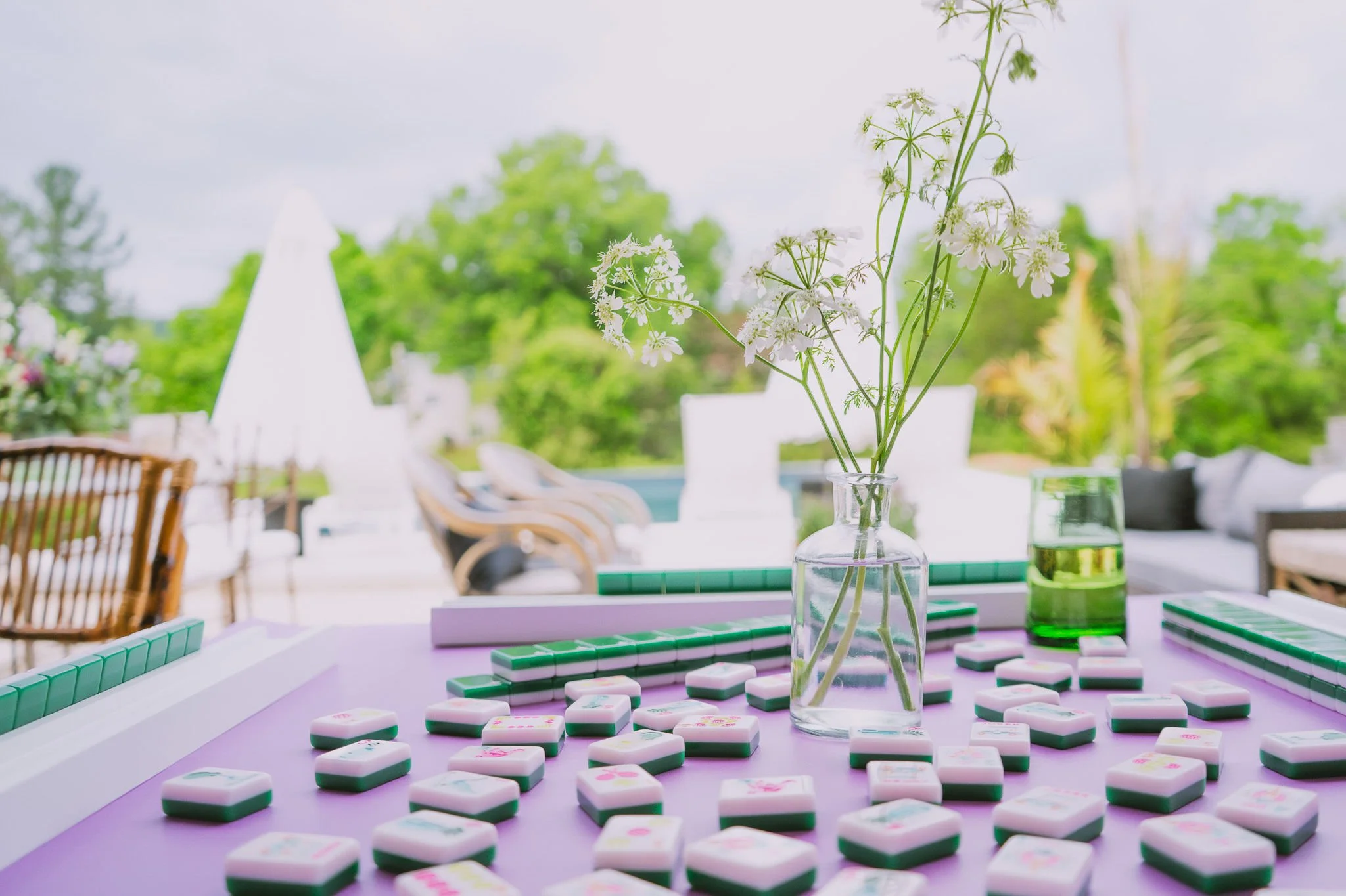 A table with Mahjong tiles scattered on its surface, a flower vase with white flowers, and outdoor patio furniture with trees in the background.