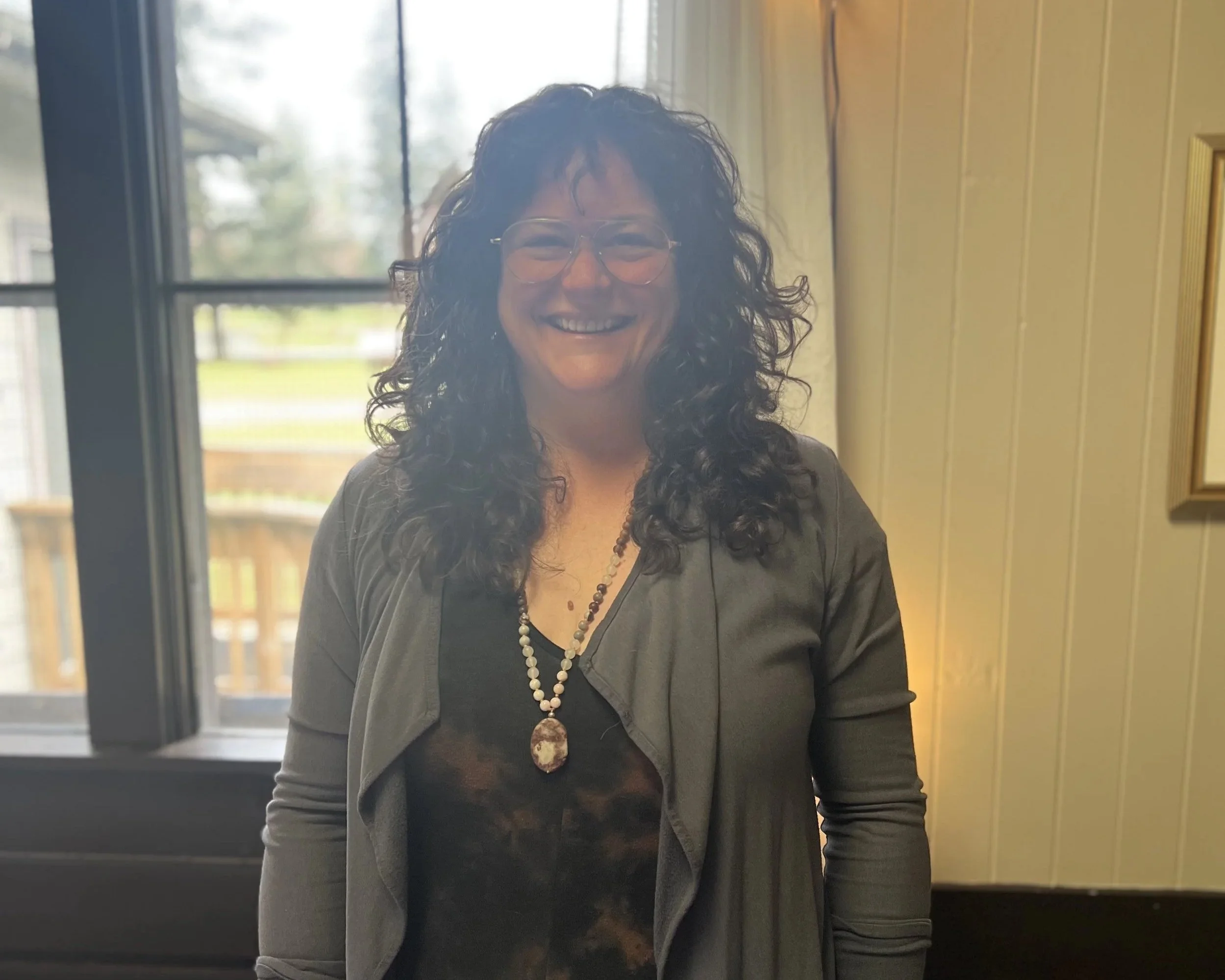 A woman with curly black hair wearing glasses, a black top, a necklace with a large pendant, and a gray jacket, smiling indoors with windows and yellow walls behind her.