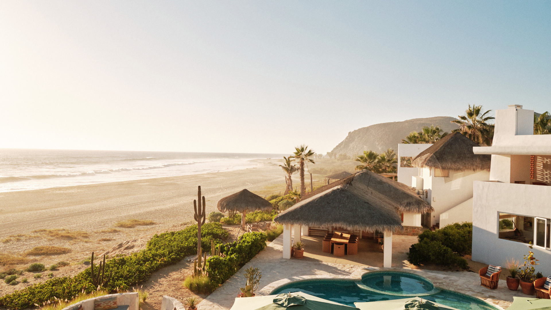 A view of a beachside resort with thatched-roof cabanas, a swimming pool, palm trees, and the ocean waves in the background on a sunny day.