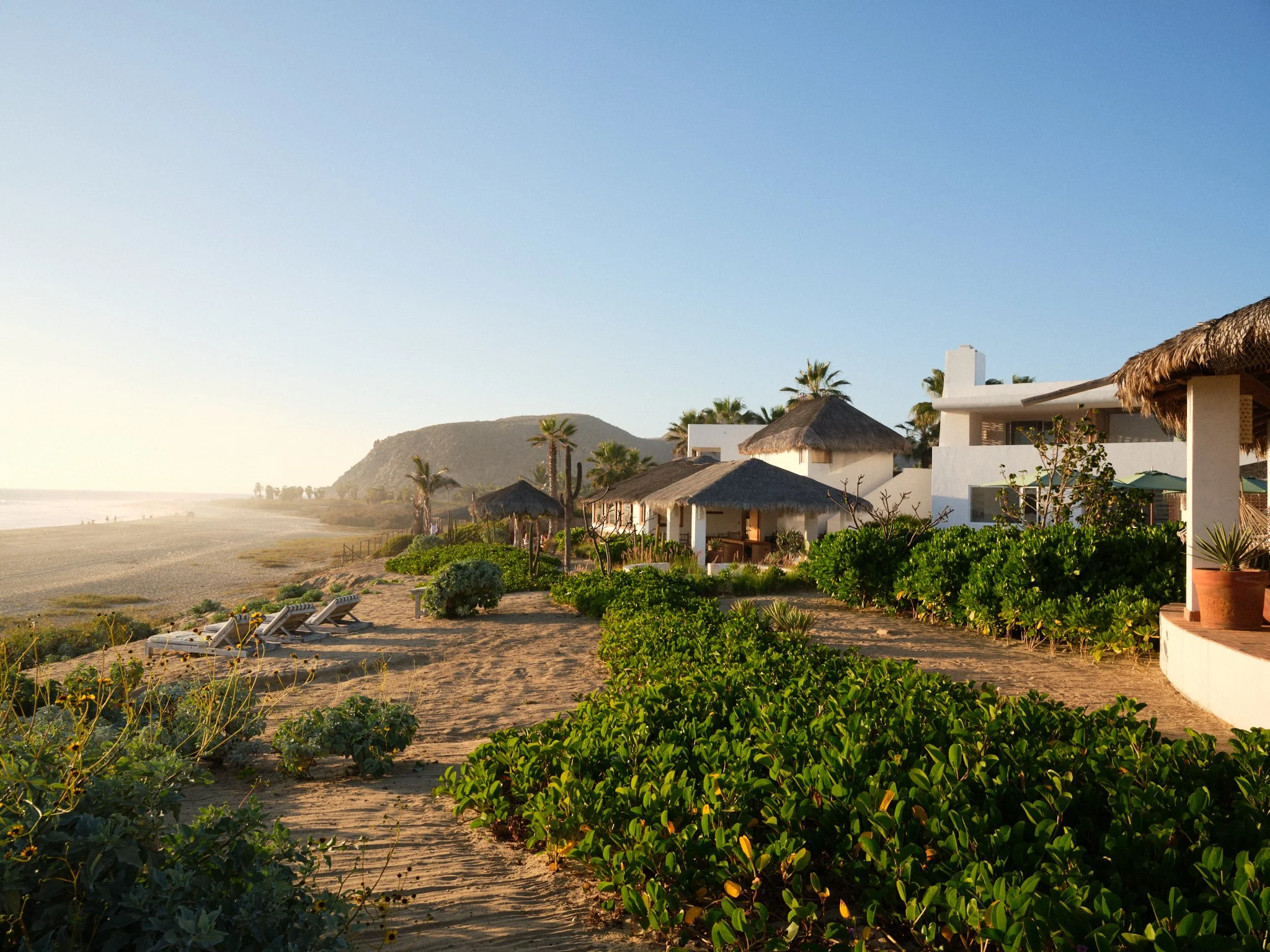 Beachside view with sandy shore, lounge chairs, lush greenery, palm trees, white modern houses with thatched roofs, and mountains in the background under a clear blue sky.