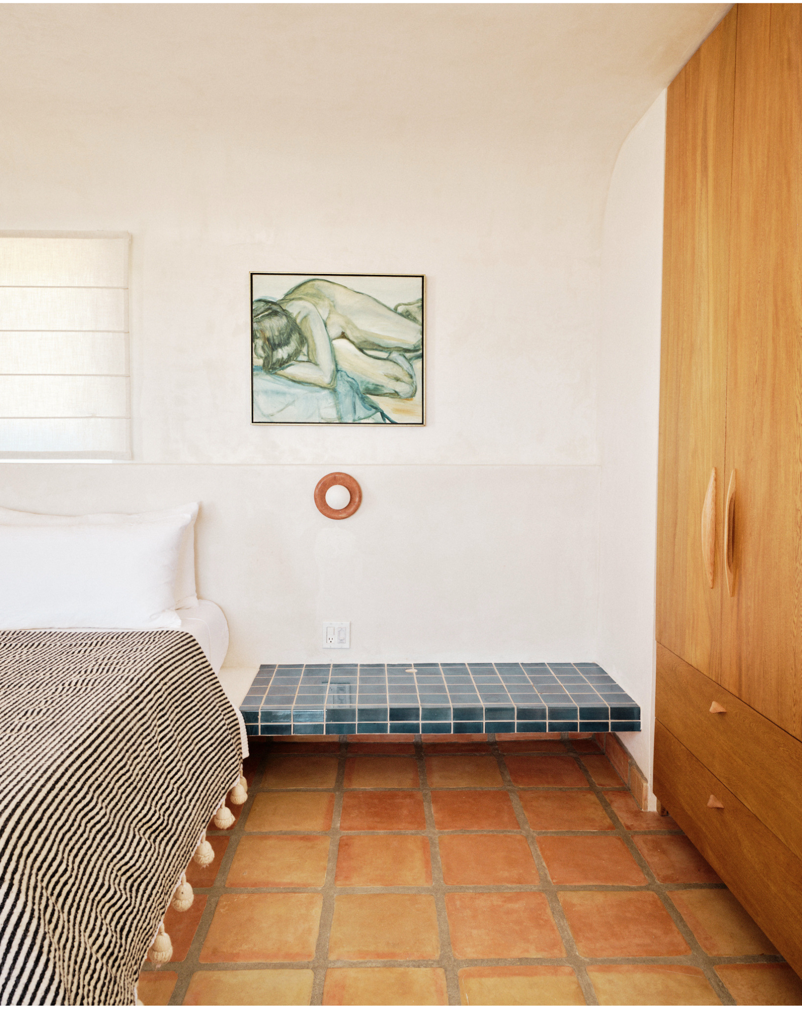 Interior view of a room with terracotta tiled floor, a white bed with a striped blanket, a wooden cabinet with rounded handles, a window with blinds, a framed painting of a reclining nude, a circular wall decoration, and a tiled platform under the painting.