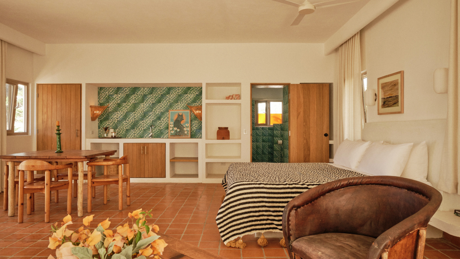 Interior view of a tropical-style hotel room with a bed, wooden furniture, decorative wall art, green tiled bathroom, and terracotta tile flooring.