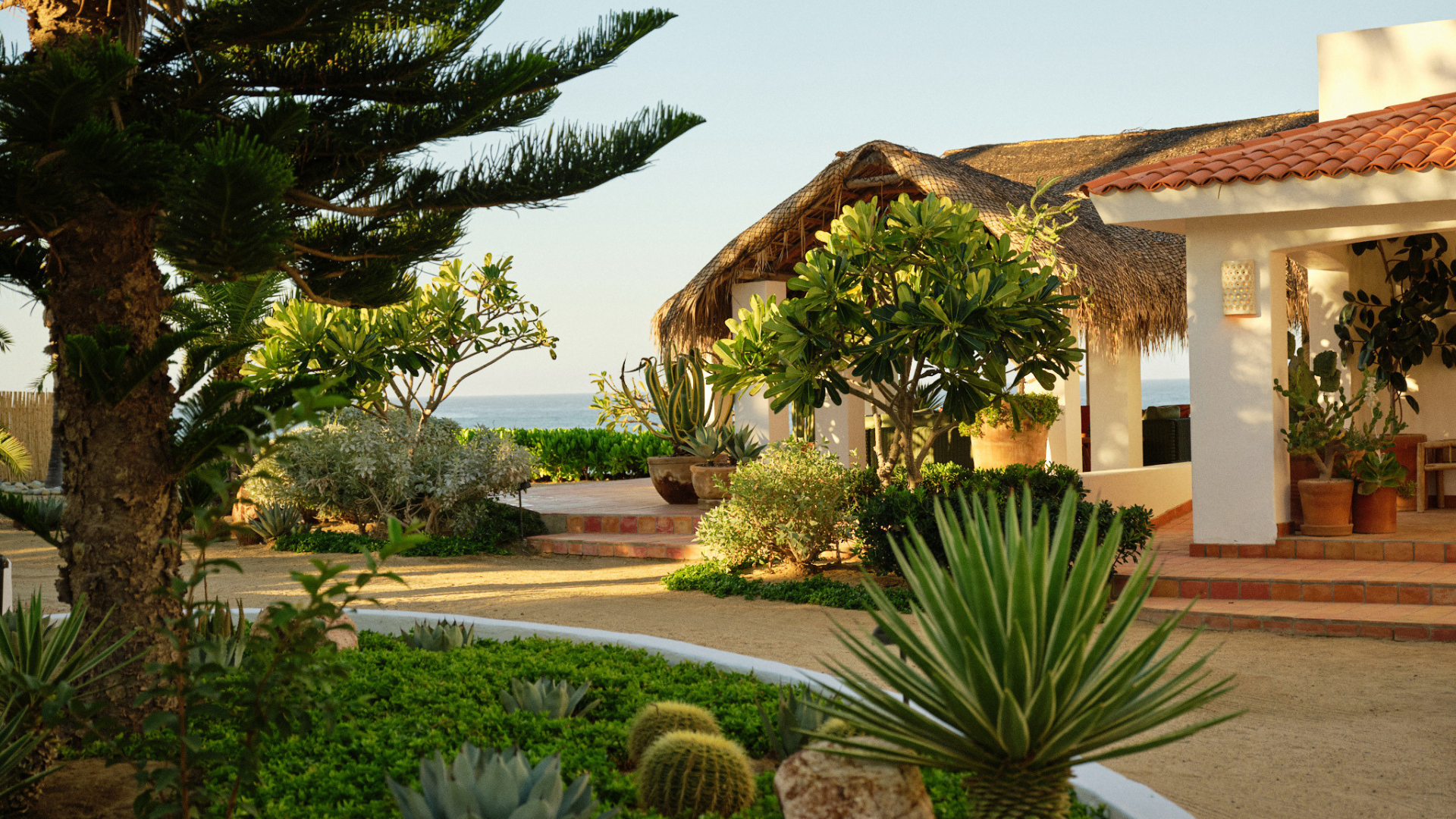 Tropical garden with various succulents and plants, white building with red tile roof, thatched roof structure, ocean in the background, sunny weather.