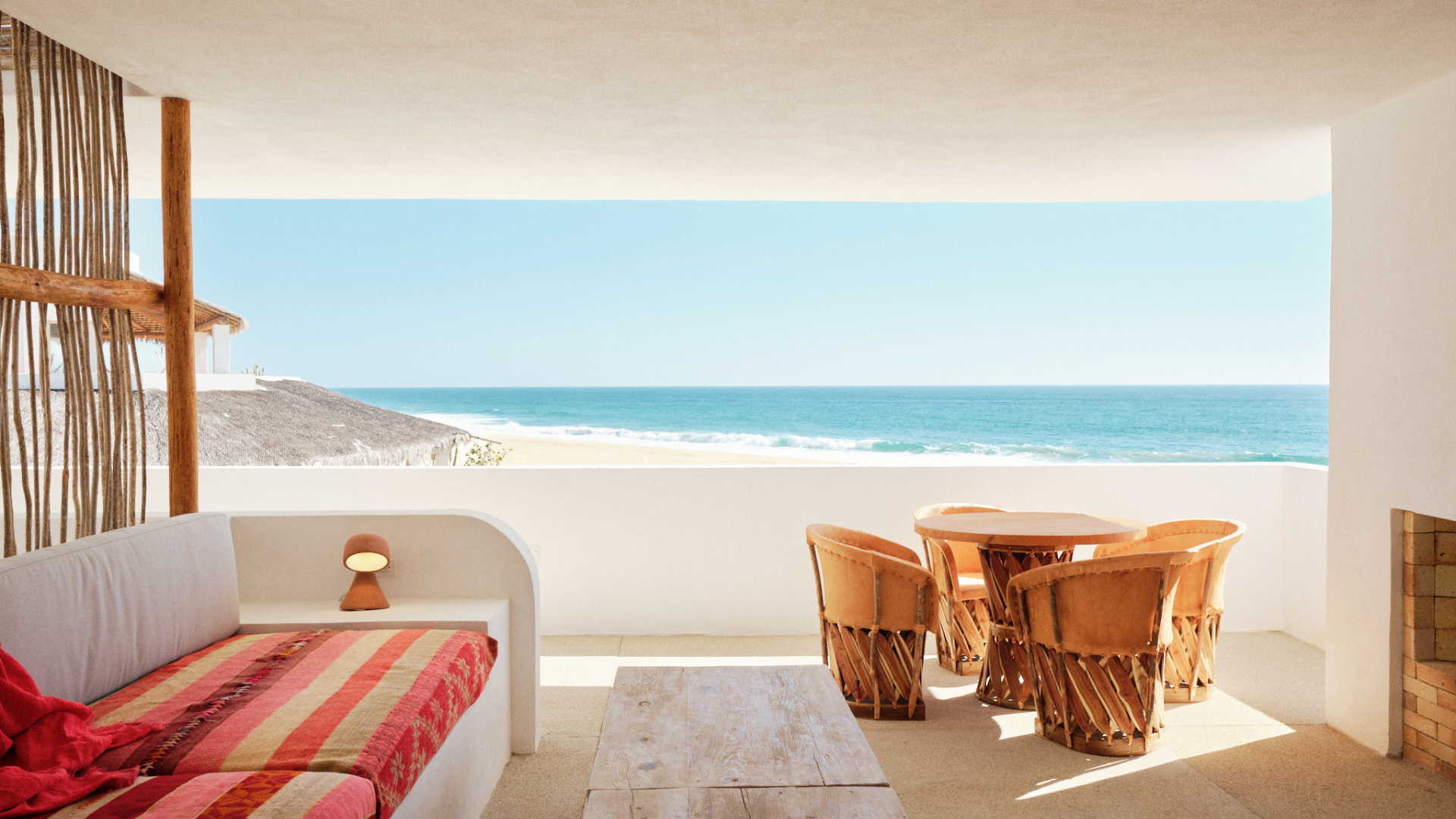 Interior view of a beachside patio with a white couch, a red and orange striped cushion, a small lamp, round wooden dining table with six chairs, and a view of the ocean and sandy beach through an open balcony.