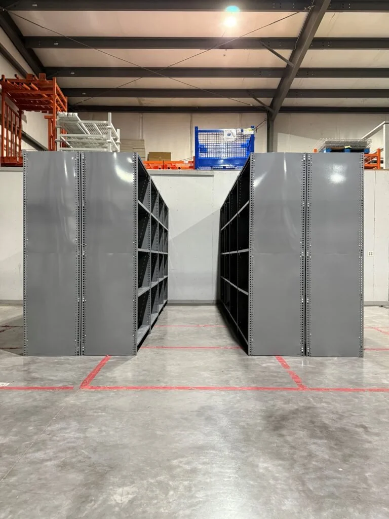 Two tall, gray industrial shelving units with multiple empty compartments in a warehouse with gray concrete floors and a high ceiling.