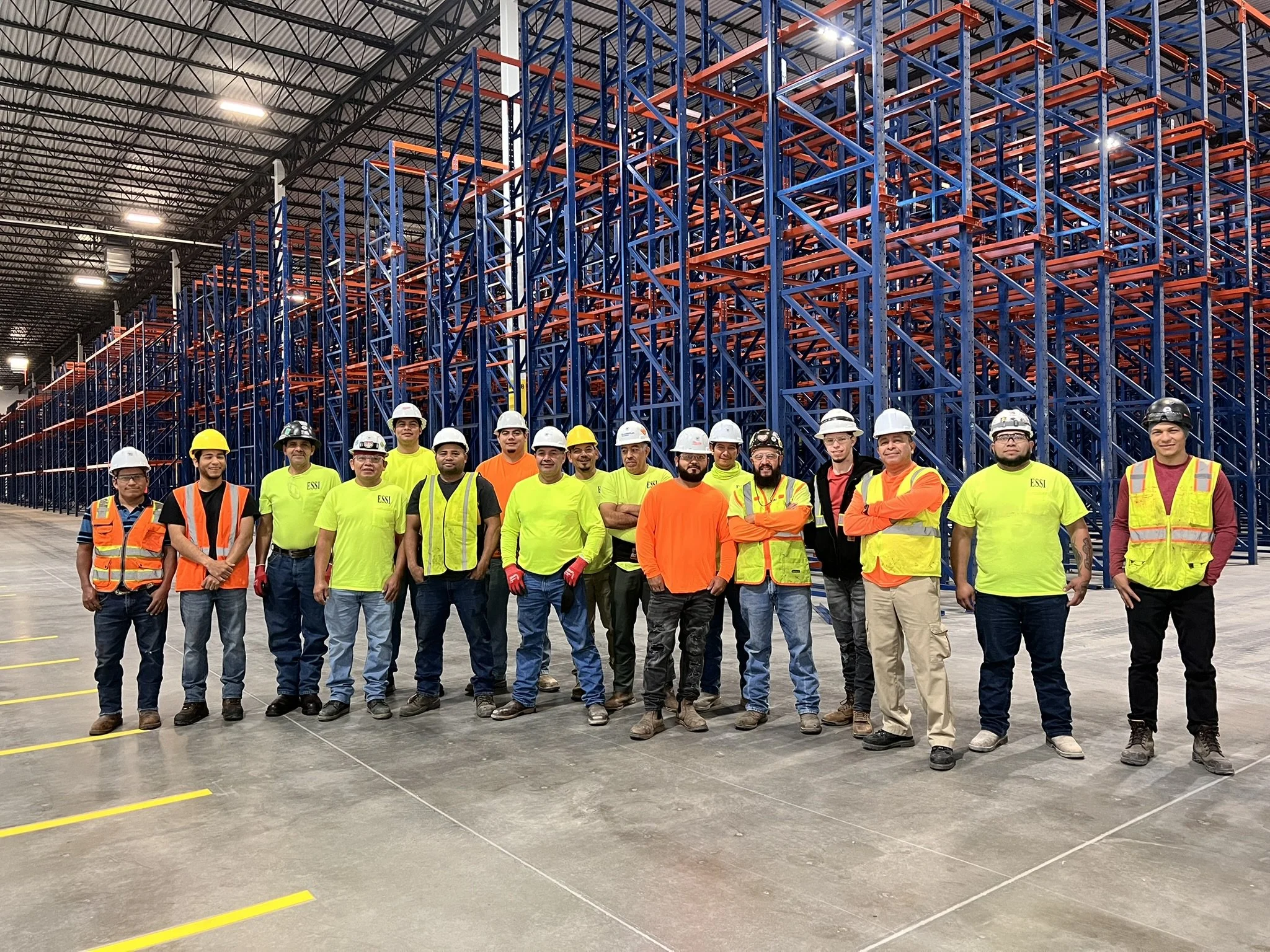A group of 15 workers standing inside a large warehouse with a high ceiling. They are wearing safety helmets and high-visibility vests. Behind them are tall blue and red metal storage racks.