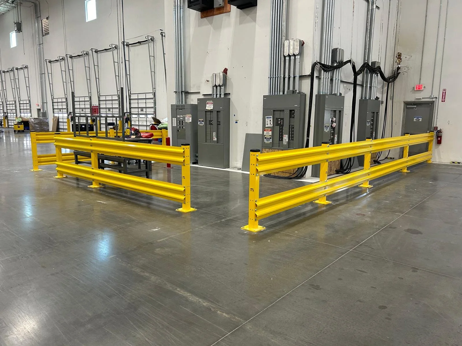 A warehouse or industrial space with four electrical control panels on a white wall, surrounded by yellow safety barriers, with some tools and equipment nearby.