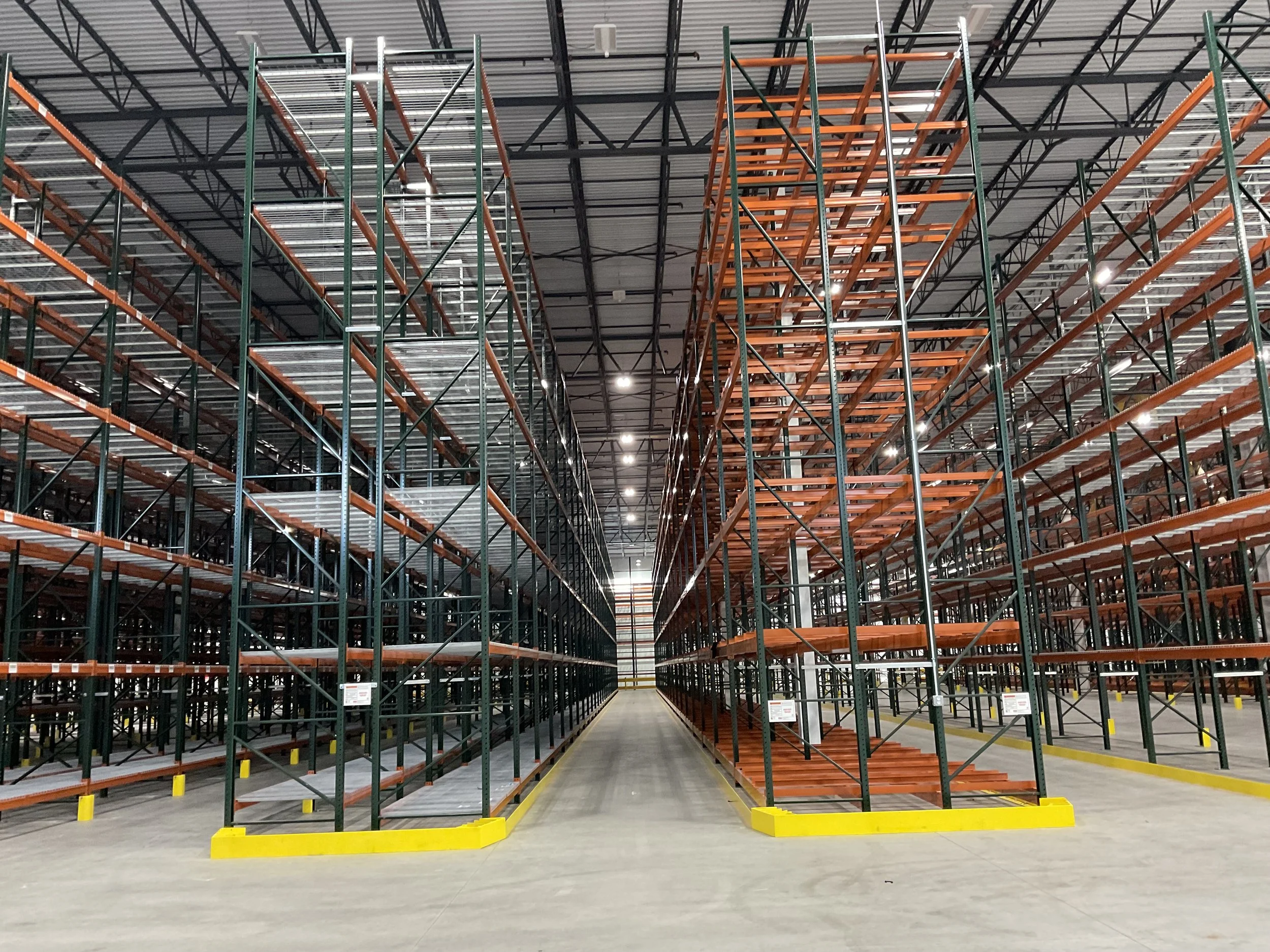Empty warehouse aisle with metal shelving units, some orange and some silver, extending upwards. The floor is polished concrete with yellow safety barriers at the front and a wide aisle running down the middle. Overhead lights illuminate the space.