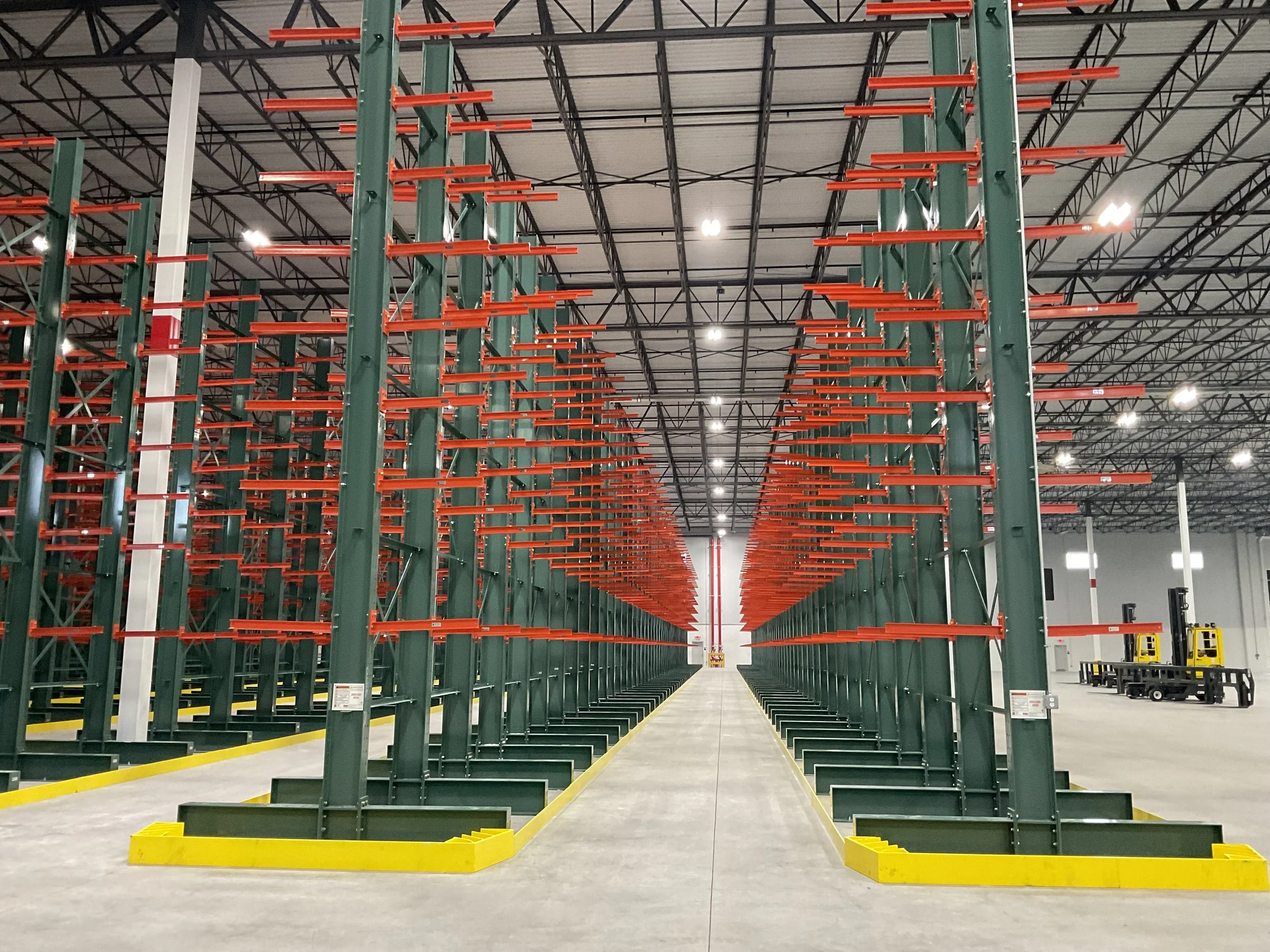Empty warehouse aisle with tall green shelving units, orange safety rails, and yellow safety barriers on the floor.