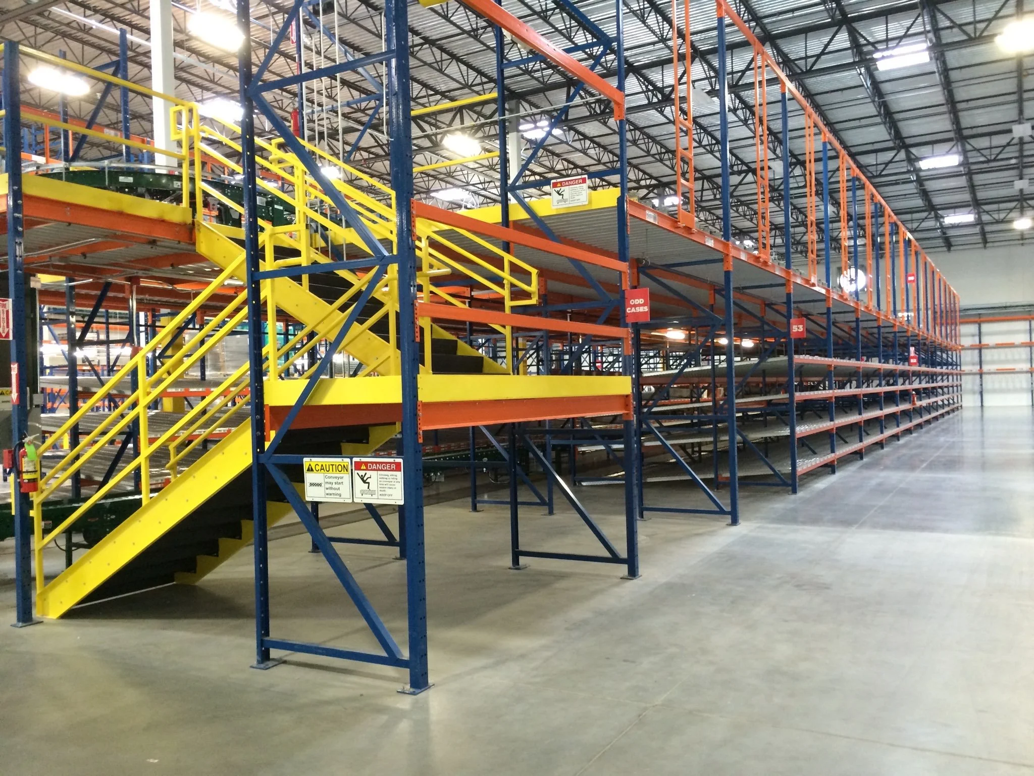 Empty warehouse aisle with blue, orange, and yellow metal storage racks, some with boxes, and yellow stairs with caution and danger signs.