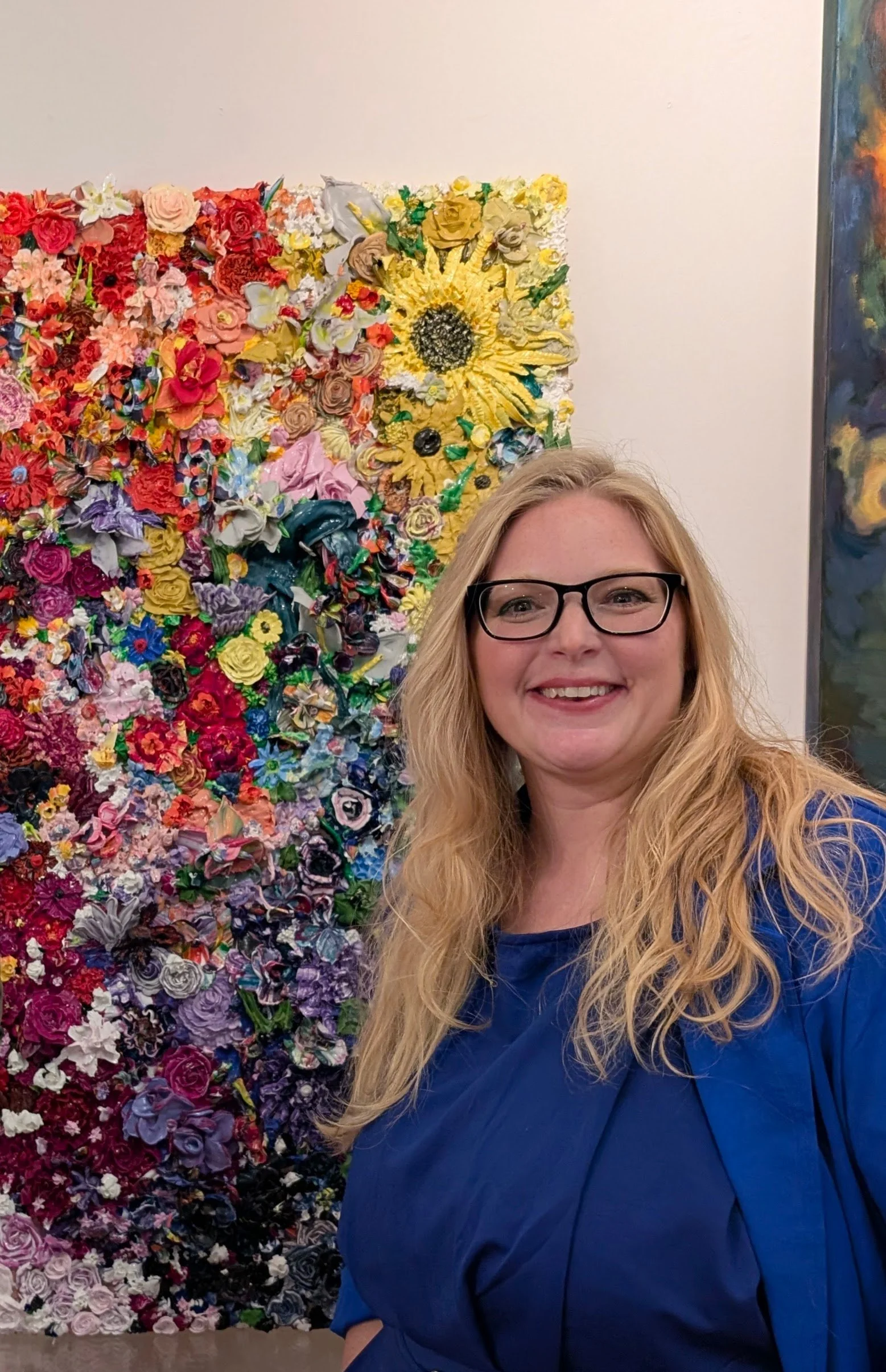 April with long blonde hair, glasses, and a blue top standing in front of a large floral wall display at an art gallery.