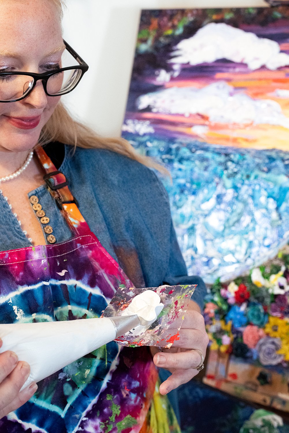 April wearing glasses and a colorful apron is using a piping bag to make a paint flower. She is standing in front of a vibrant abstract painting and a floral arrangement.