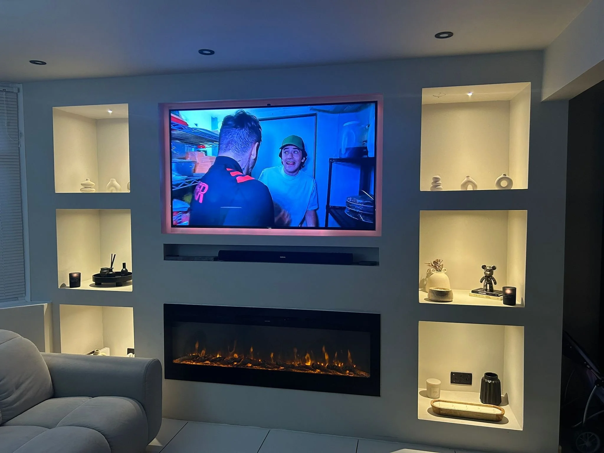 Living room with a large wall-mounted television, The room features a media wall with built-in shelves with decorative items, a fireplace below the TV with a glowing fire, a grey sofa in the foreground, and various small objects on surfaces.