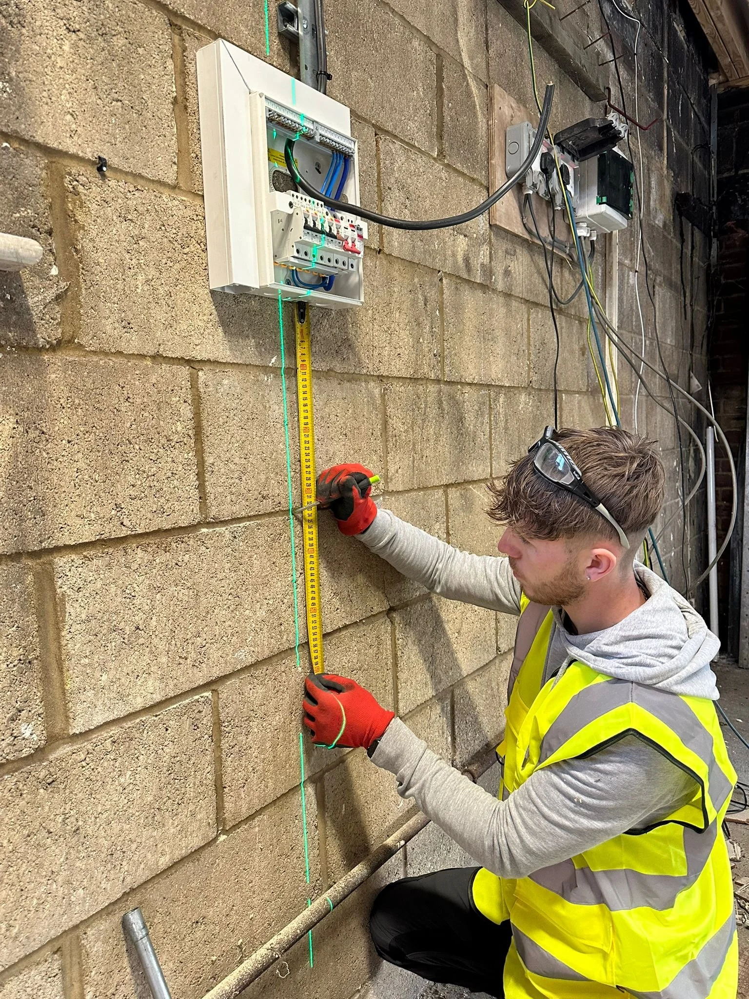 A worker in a yellow safety vest measuring a wall with a tape measure. The worker is wearing red gloves and sunglasses, and is working near an electrical box mounted on a brick wall. Green laser lines are projected on the wall for alignment.