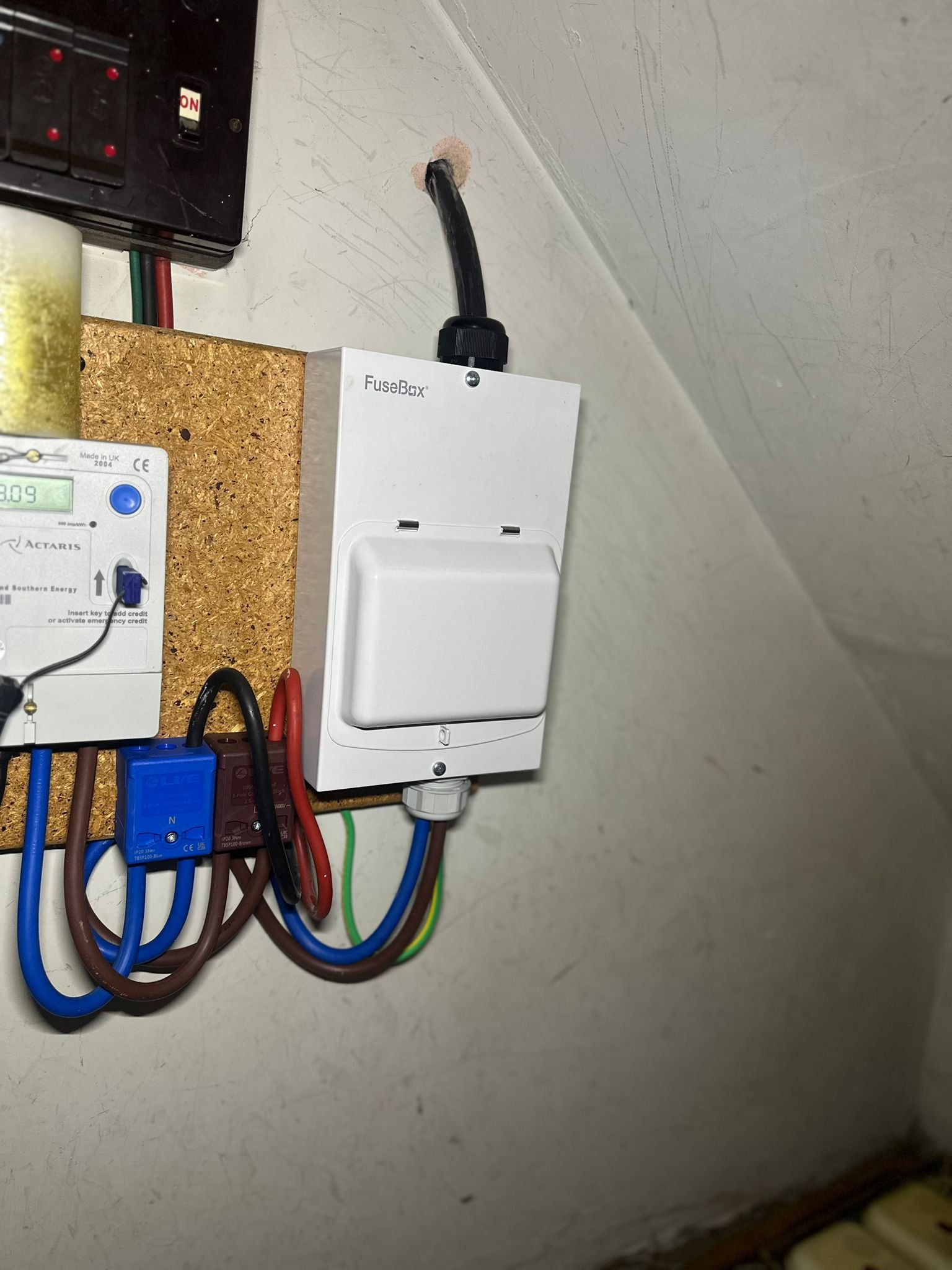 Electrical fusebox with circuit breakers and wiring, mounted on a wall with a textured cork board beneath and a dno meter.