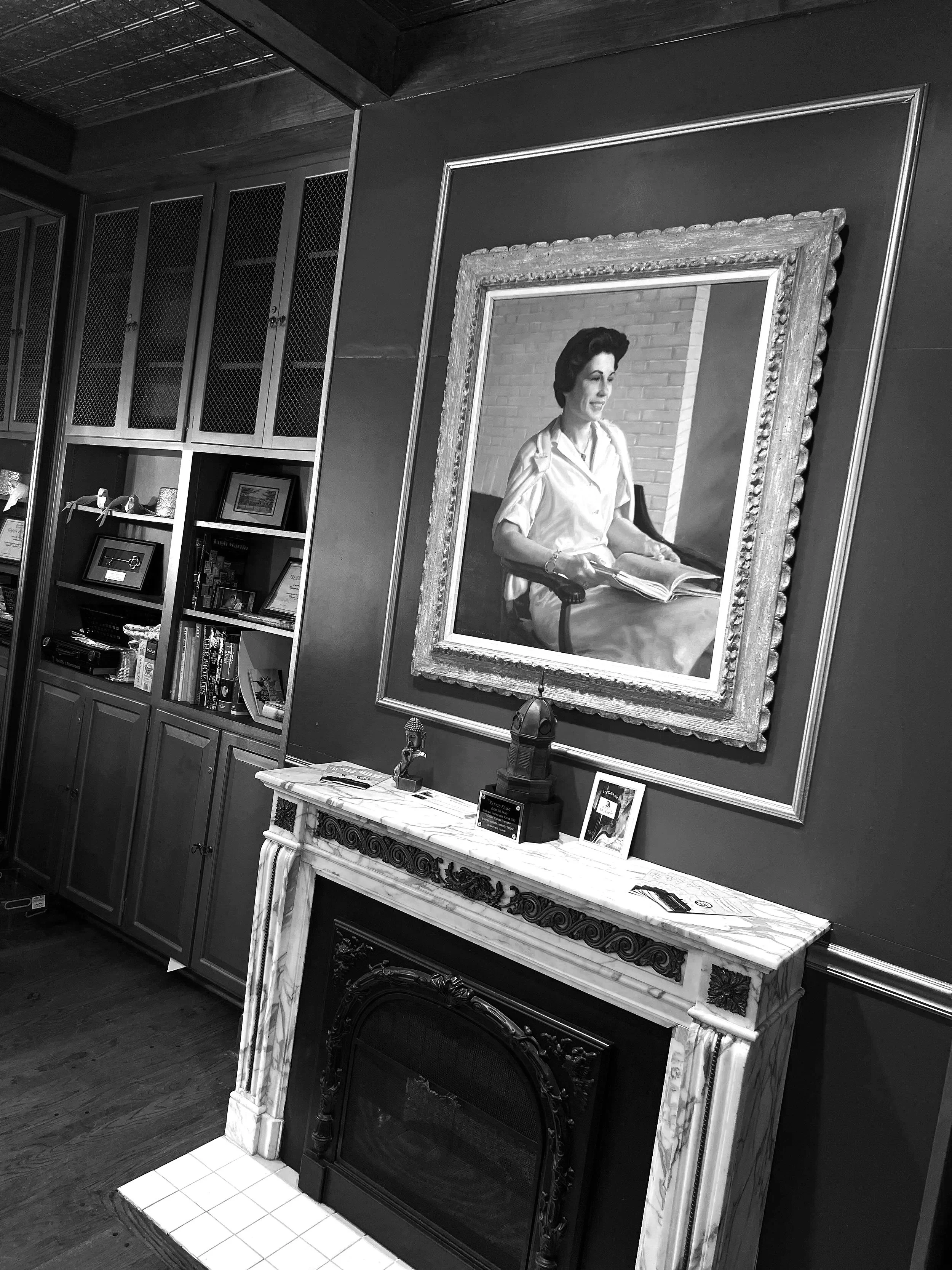 A black and white photo of a room with a large ornate portrait mirror on a wall, a decorative fireplace with a marble mantel, a small table with trophies and picture frames, and built-in bookshelves filled with books and framed documents.