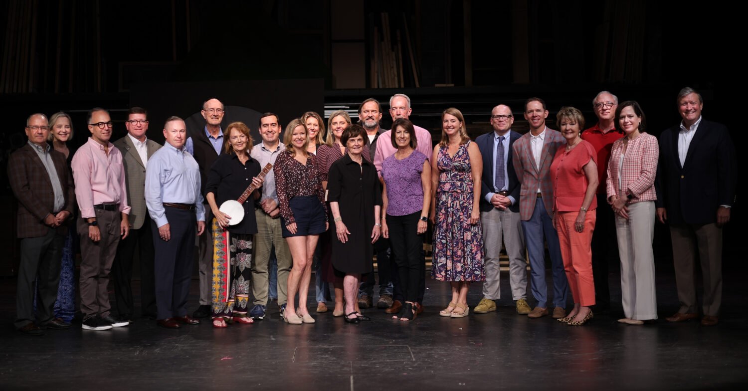 Group of people standing on stage for a portrait, some smiling, with wooden floors and dark background.