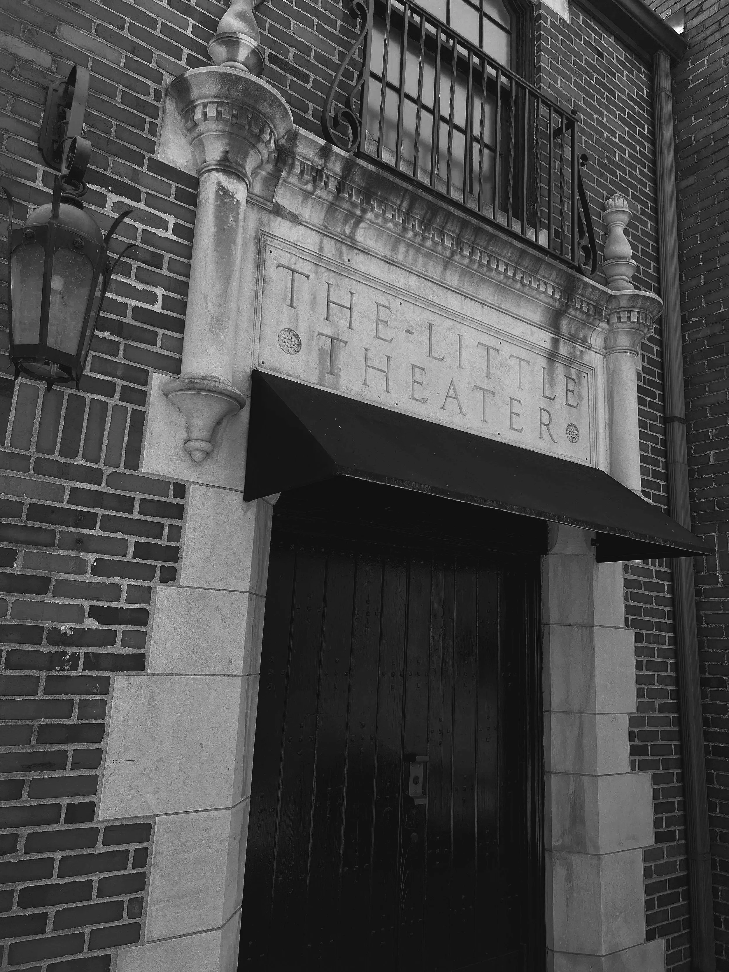 The entrance to a theater with a stone sign that reads 'The Little Theatre,' decorative stone columns, a hanging lantern, a metal railing above the sign, and brick walls surrounding the entrance.