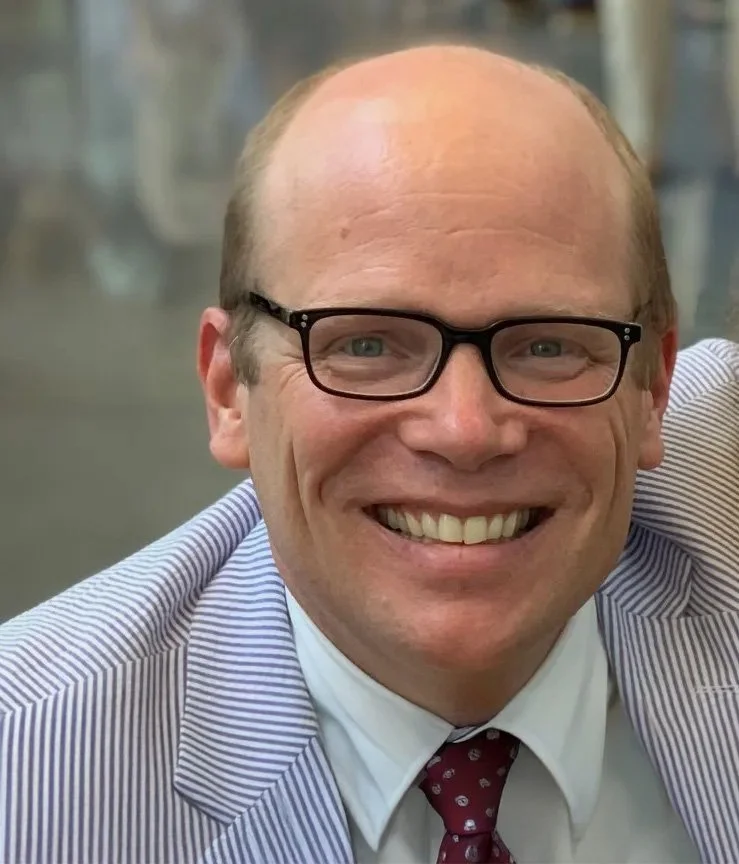 A smiling man wearing glasses, a blue and white striped suit jacket, and a burgundy tie with white dots.