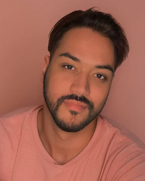 A young man with dark hair and a beard, wearing a pink shirt, taking a selfie against a pink background.