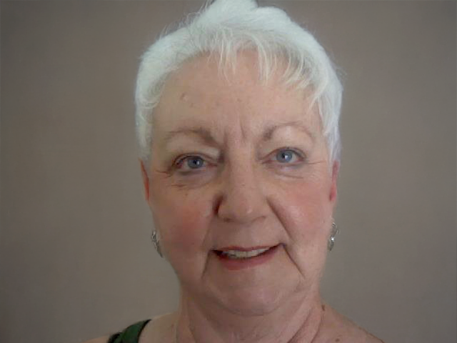 Close-up of an elderly woman with short white hair, blue eyes, and earrings, smiling slightly against a plain gray background.