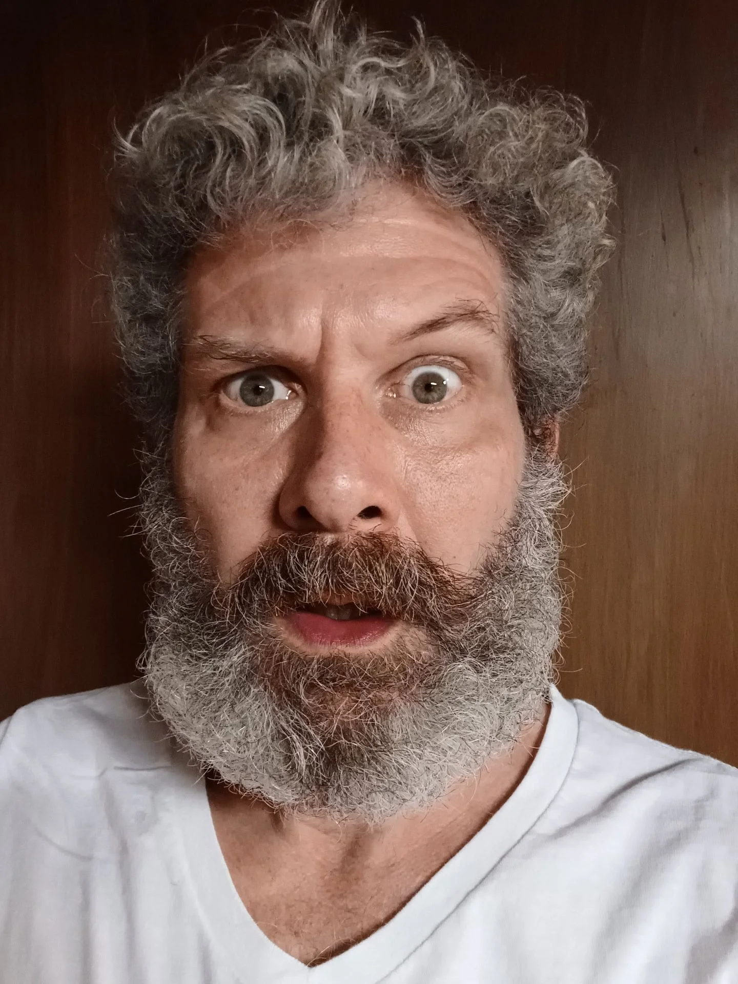 A man with curly gray hair and a full gray beard looking surprised or excited, wearing a white shirt, against a wooden background.