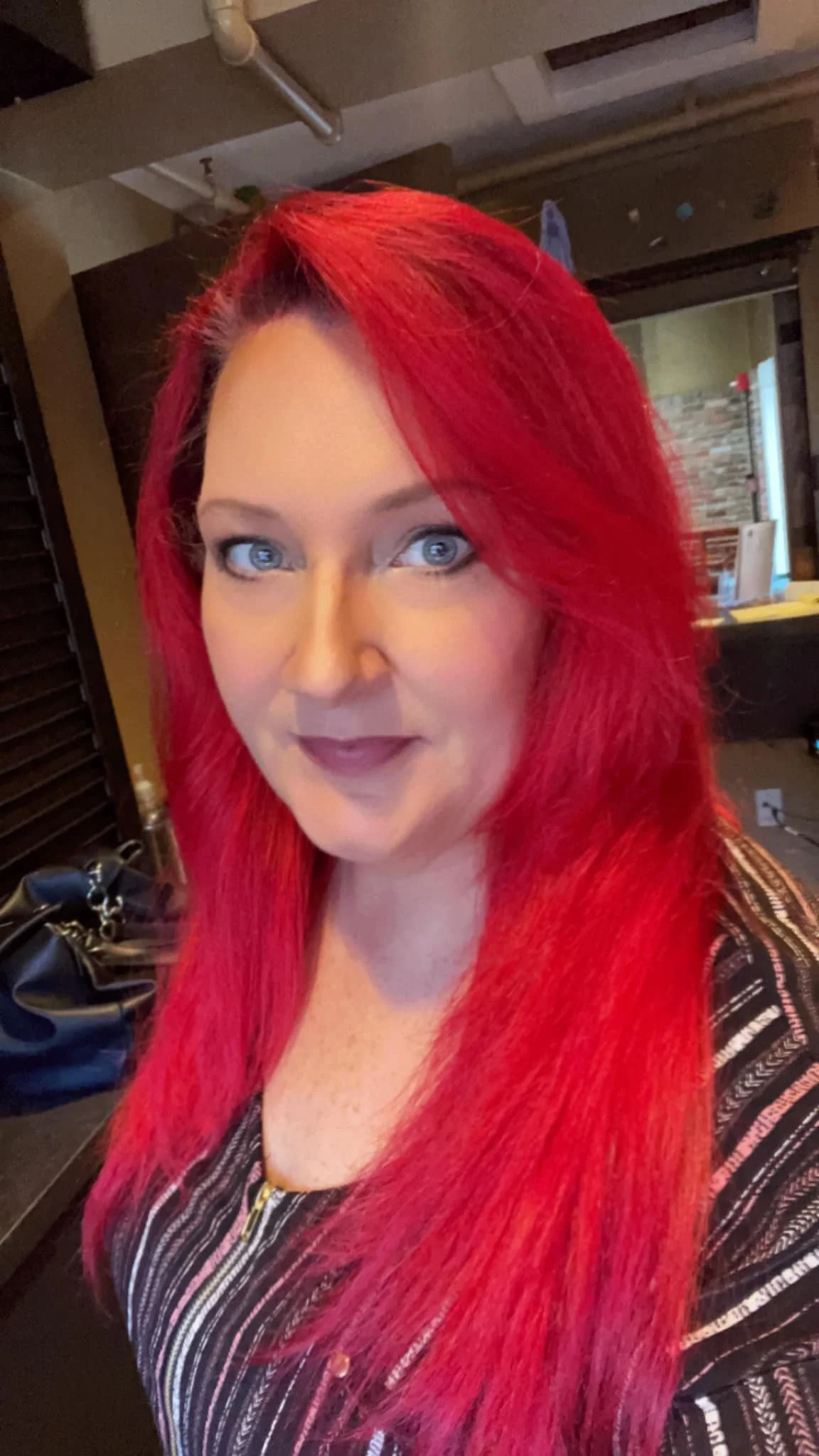 A woman with bright red hair and blue eyes taking a selfie in an indoor setting. She is wearing a striped top with black, white, and pink colors.