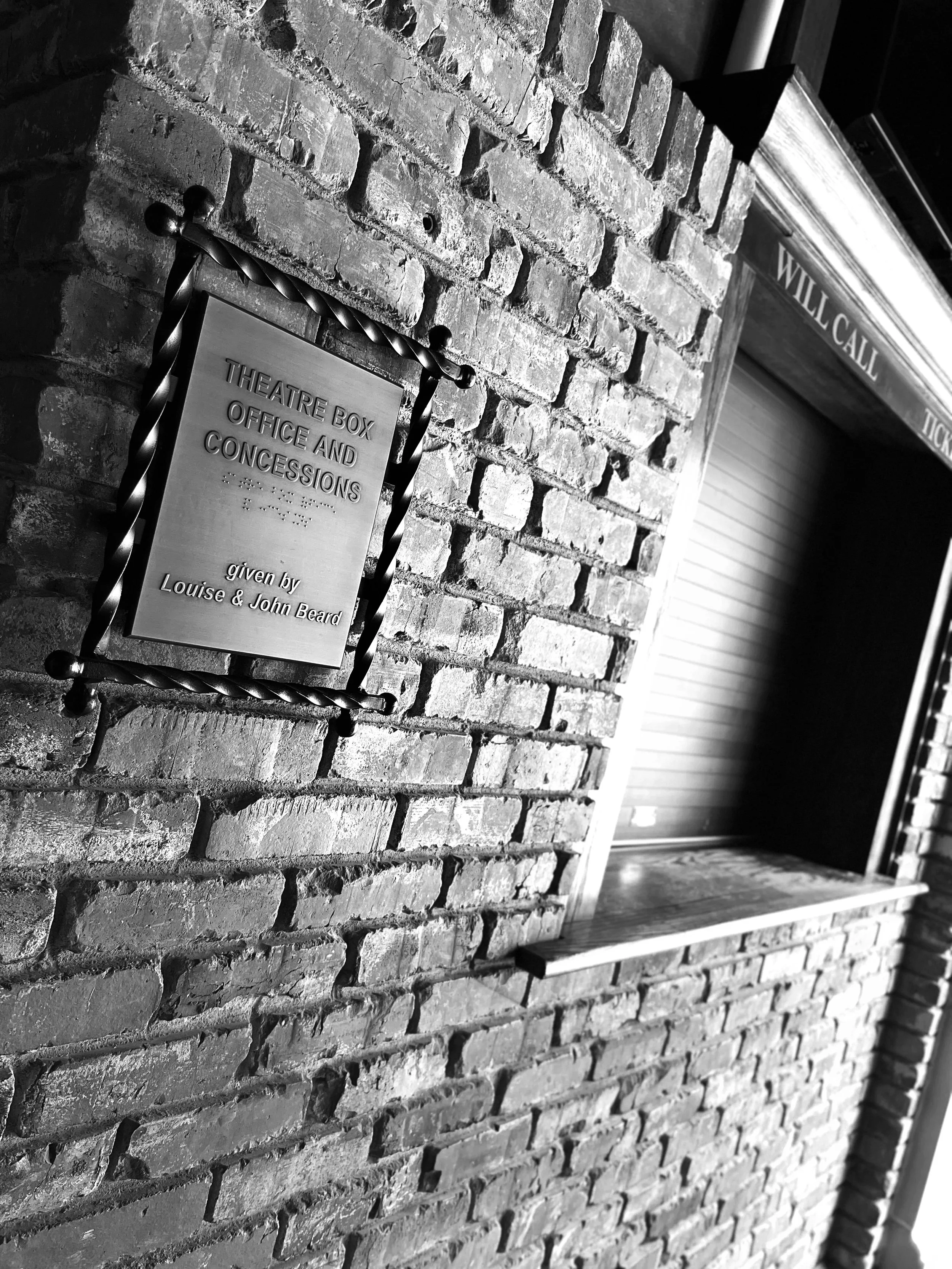 A brick wall with a metal plaque that reads 'Theatre Box Office and Concessions given by Louise & John Beard' in front of a structure, possibly a theater, with a sign that says 'Will Call Tickets' in the background.
