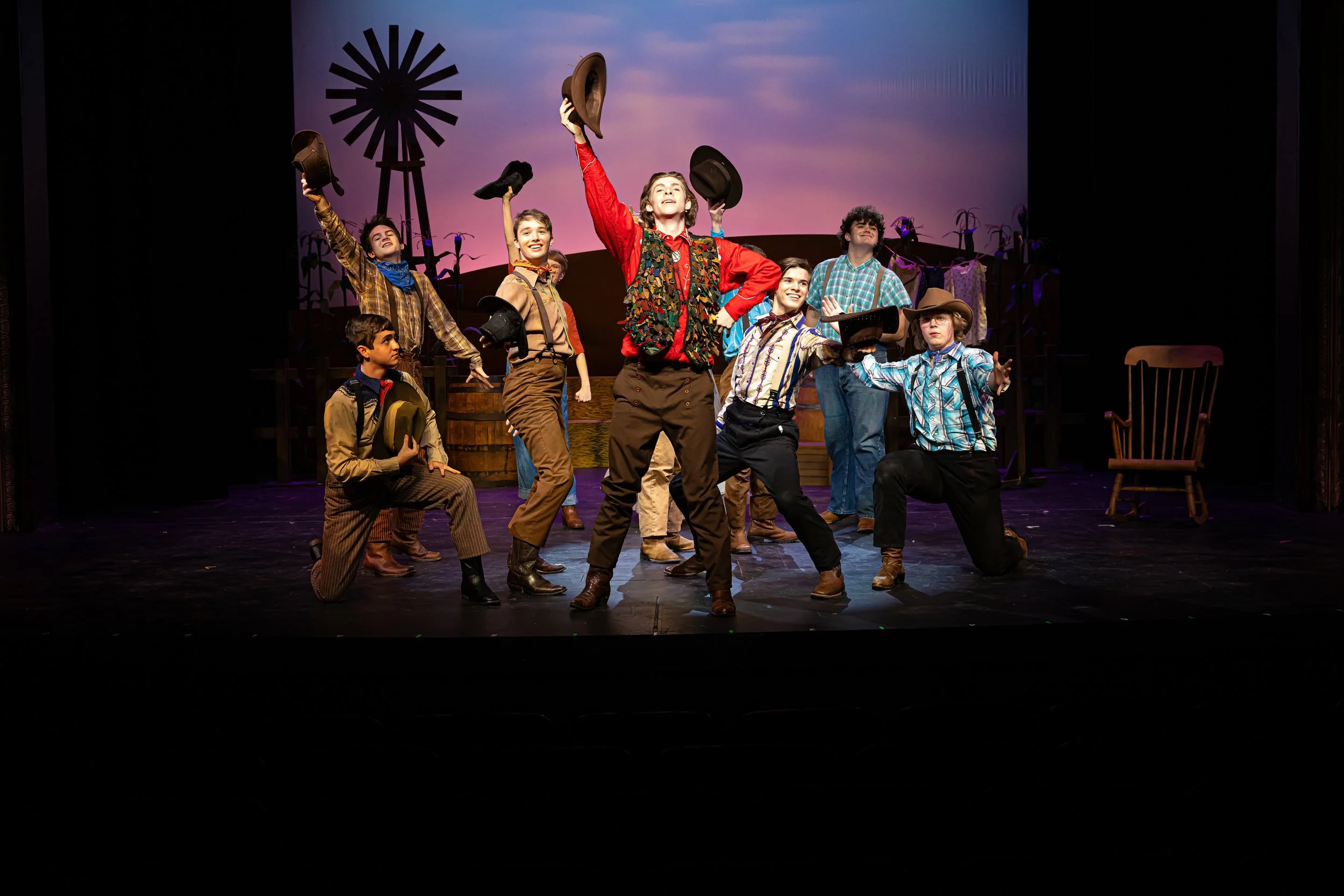 Group of children dressed in cowboy and cowgirl costumes, standing on a stage with scenic backdrop, some holding hats, in a theater.