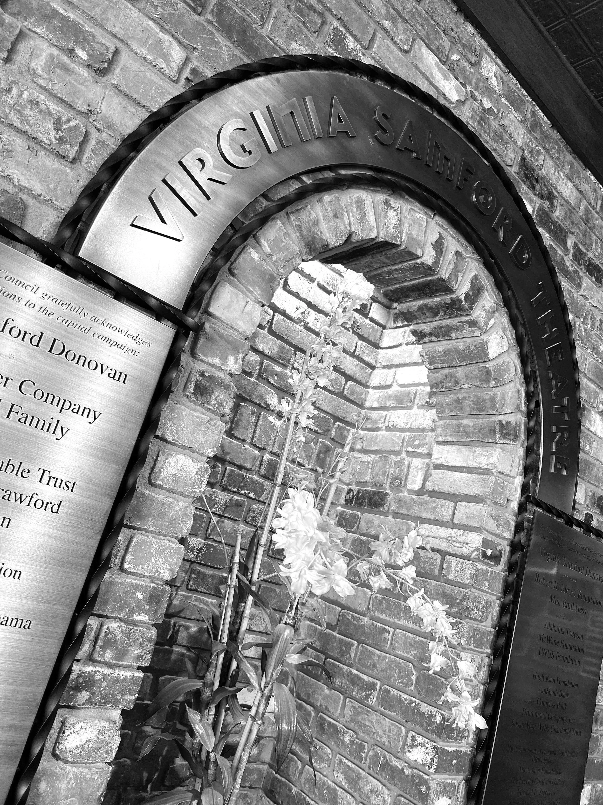 A memorial plaque mounted on a brick wall with an arched opening, featuring the words 'VIRGINIA SANDERS'. There are plants with white flowers in front of the plaque.