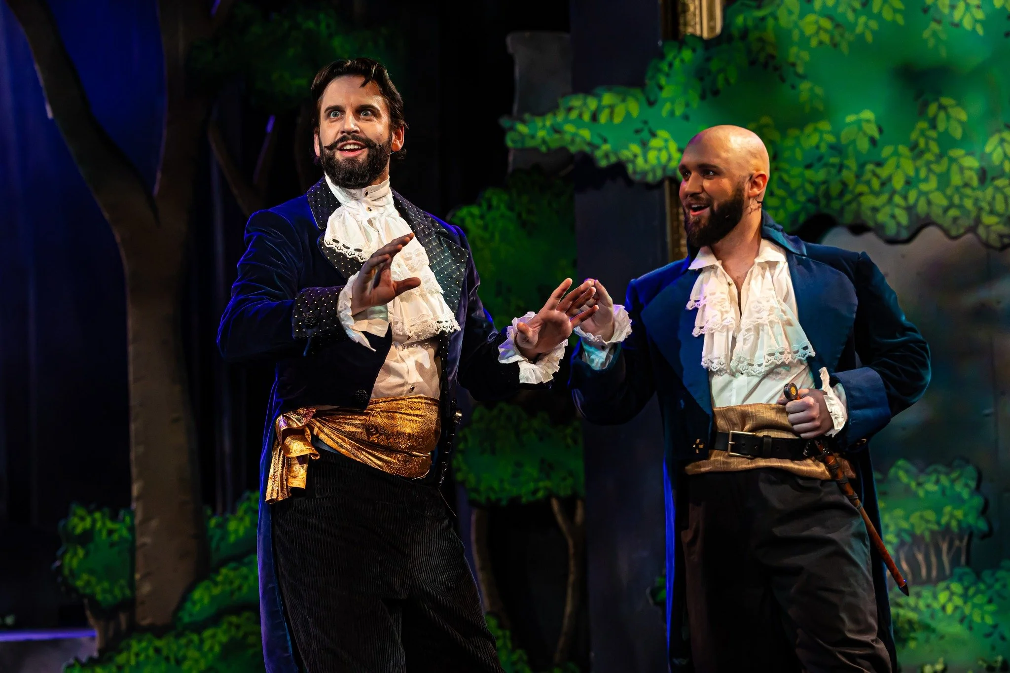 Two men performing on stage in theatrical costumes; one with a beard and vintage suit with lace collar, the other is bald with a beard in period attire, set against a backdrop of painted green leaves.