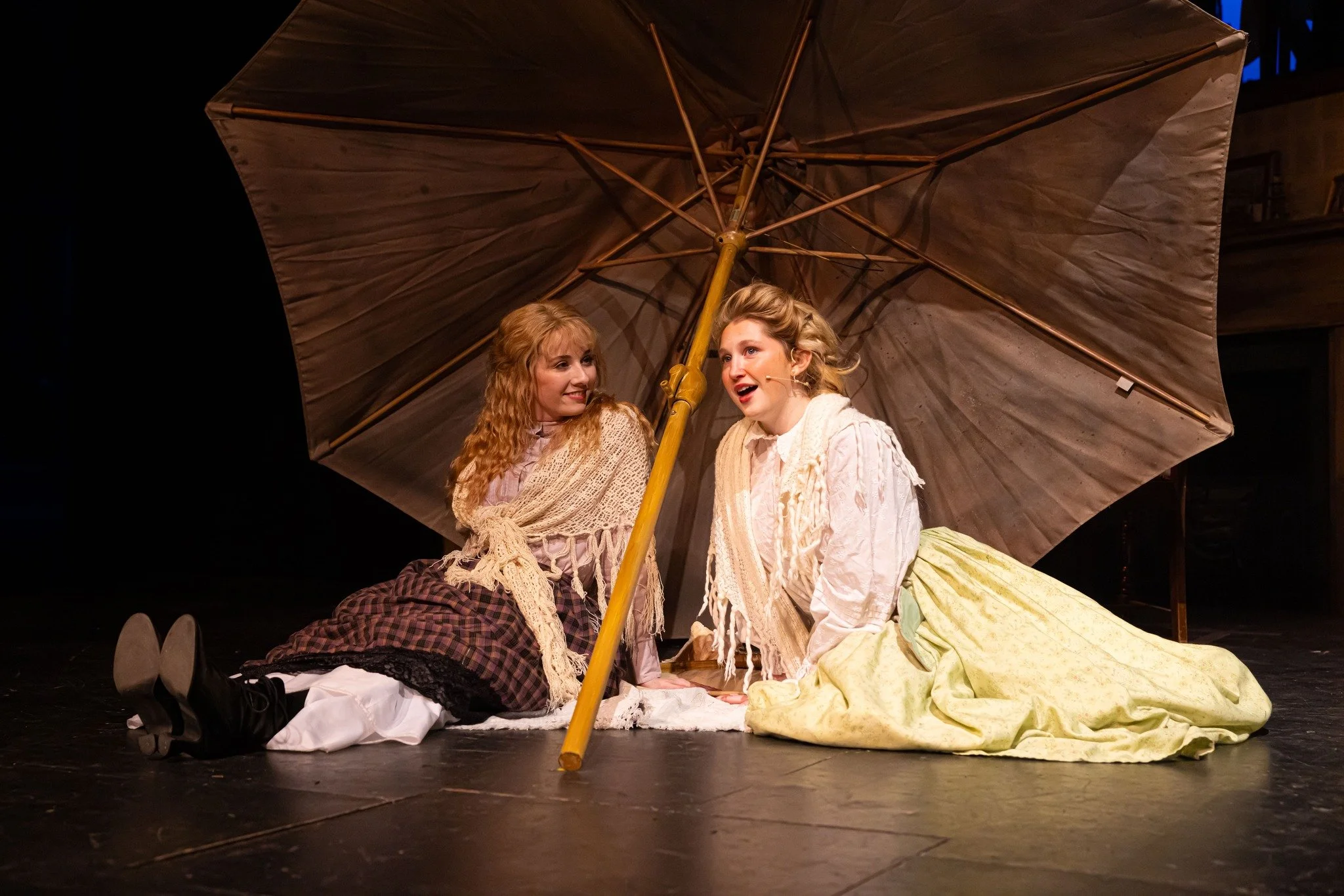 Two women sitting on stage under a large umbrella, dressed in vintage clothing, with one woman looking at the other as they perform a theatrical scene.