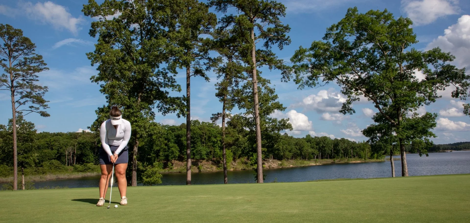 A person in golf attire preparing to putt on a golf green near a lake, surrounded by trees and a blue sky with clouds.