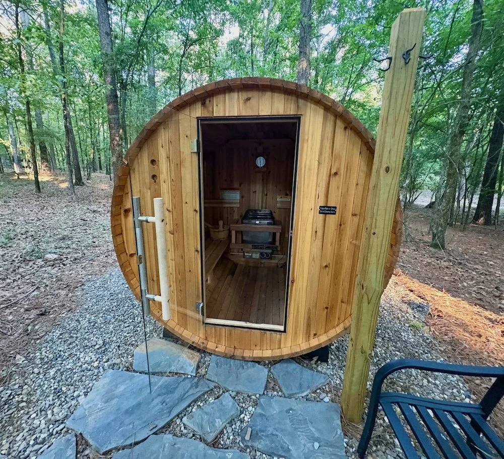 A small, round, wooden sauna with a glass door is situated in a wooded area, with stone paving leading up to it.