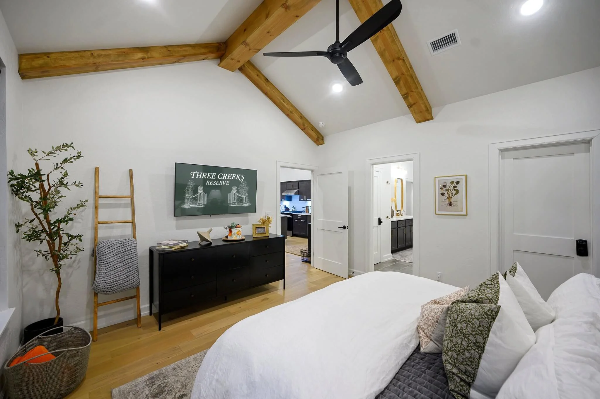 A bedroom with white walls and wooden ceiling beams. There is a bed with white bedding and decorative pillows. A black dresser with decorative items and a television is against the wall, with a framed art piece above it. There are doorway openings le