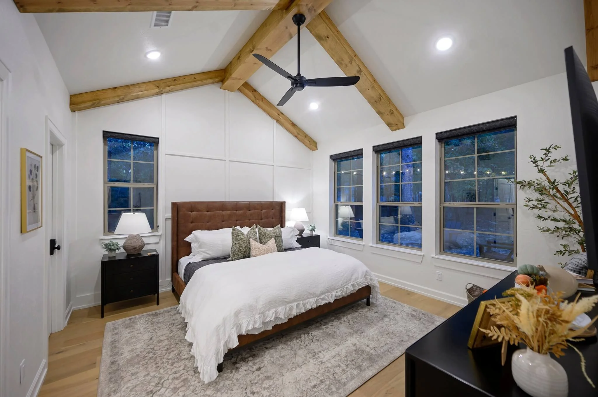 A bedroom with a high ceiling featuring exposed wooden beams and a black ceiling fan. The room has white walls with decorative molding, three windows with dark blinds, and a large bed with a wooden headboard, white bedding, and multiple pillows. Ther